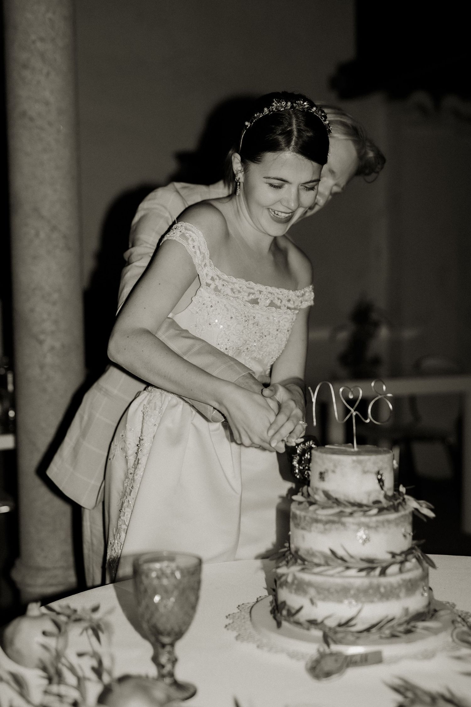 Black and white photo of smiling bride cutting three-tier wedding cake at reception.