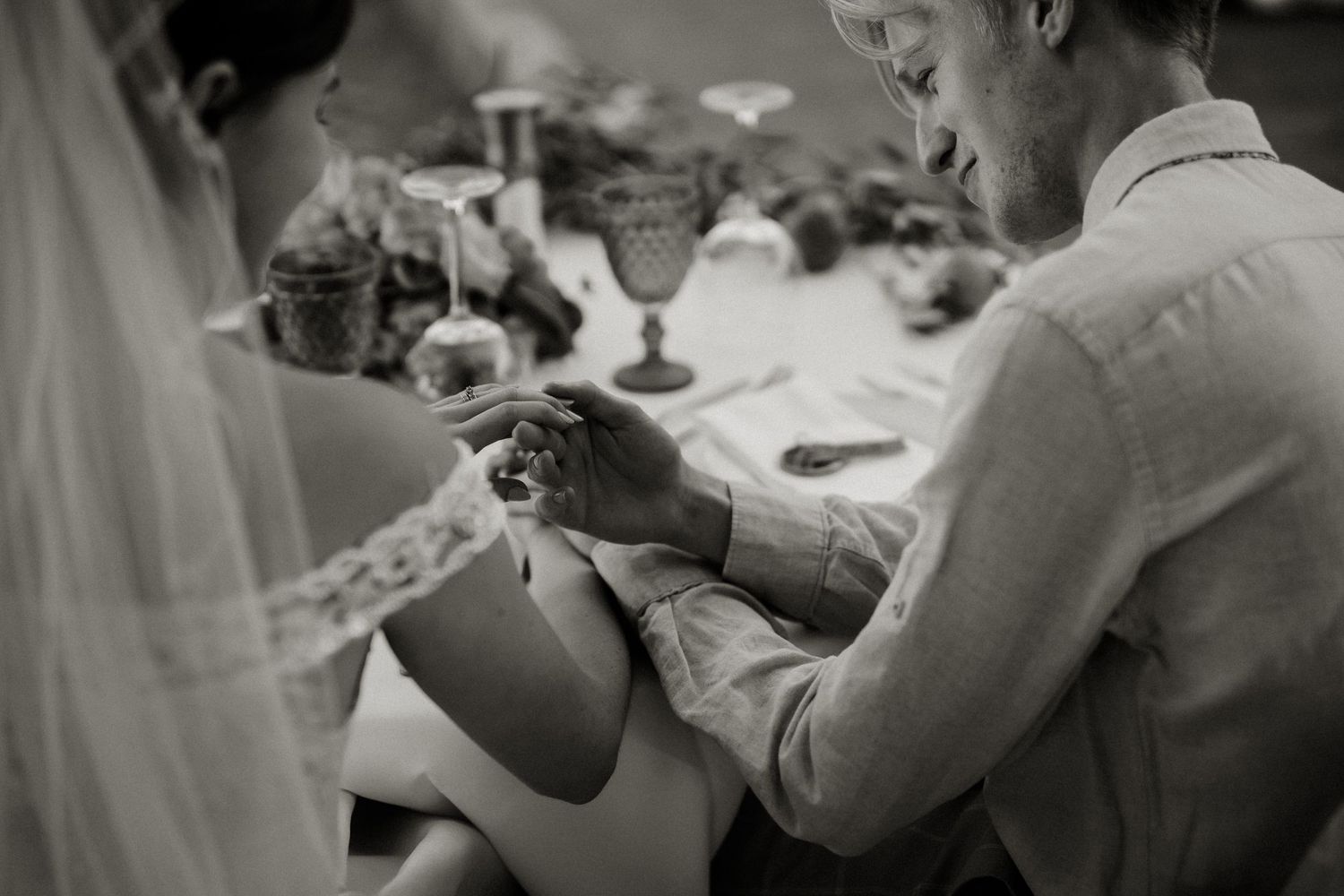Black and white photo of a groom placing a wedding ring on his bride's finger at the reception table.