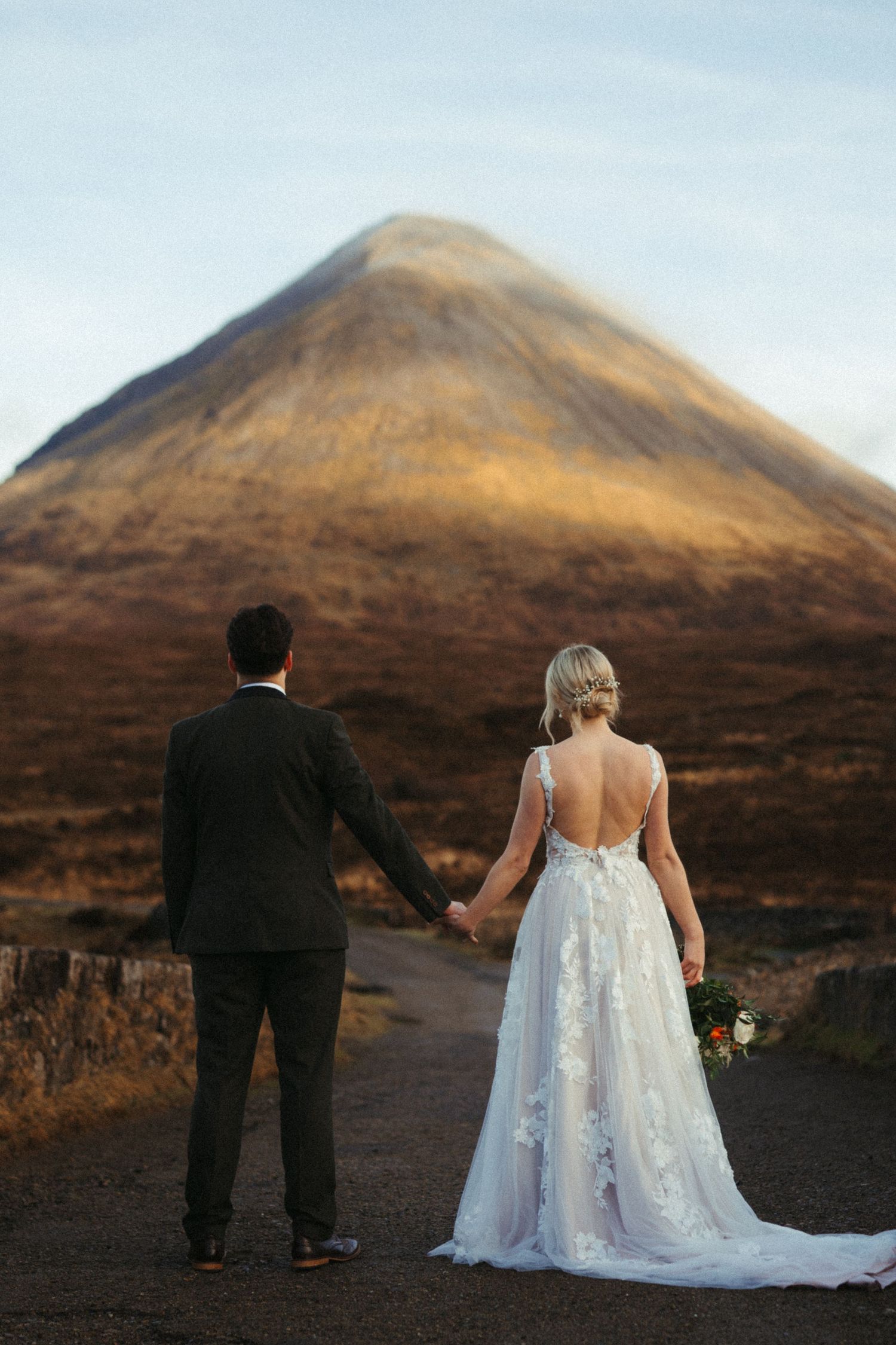 Wedding couple holding hands facing a dramatic mountain landscape at golden hour.