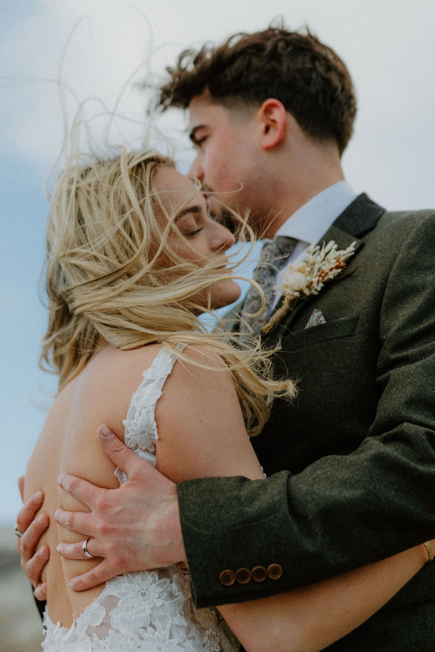Groom kisses bride as wind blows her blonde hair, both embracing in an intimate outdoor moment.
