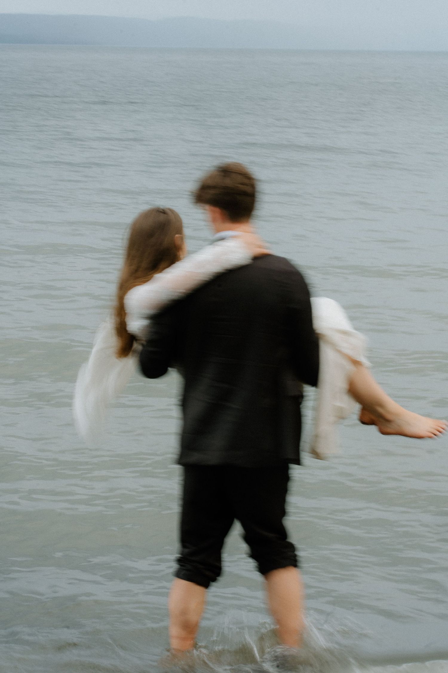 Blurred artistic shot of groom carrying bride into ocean waves for dreamy engagement photo.