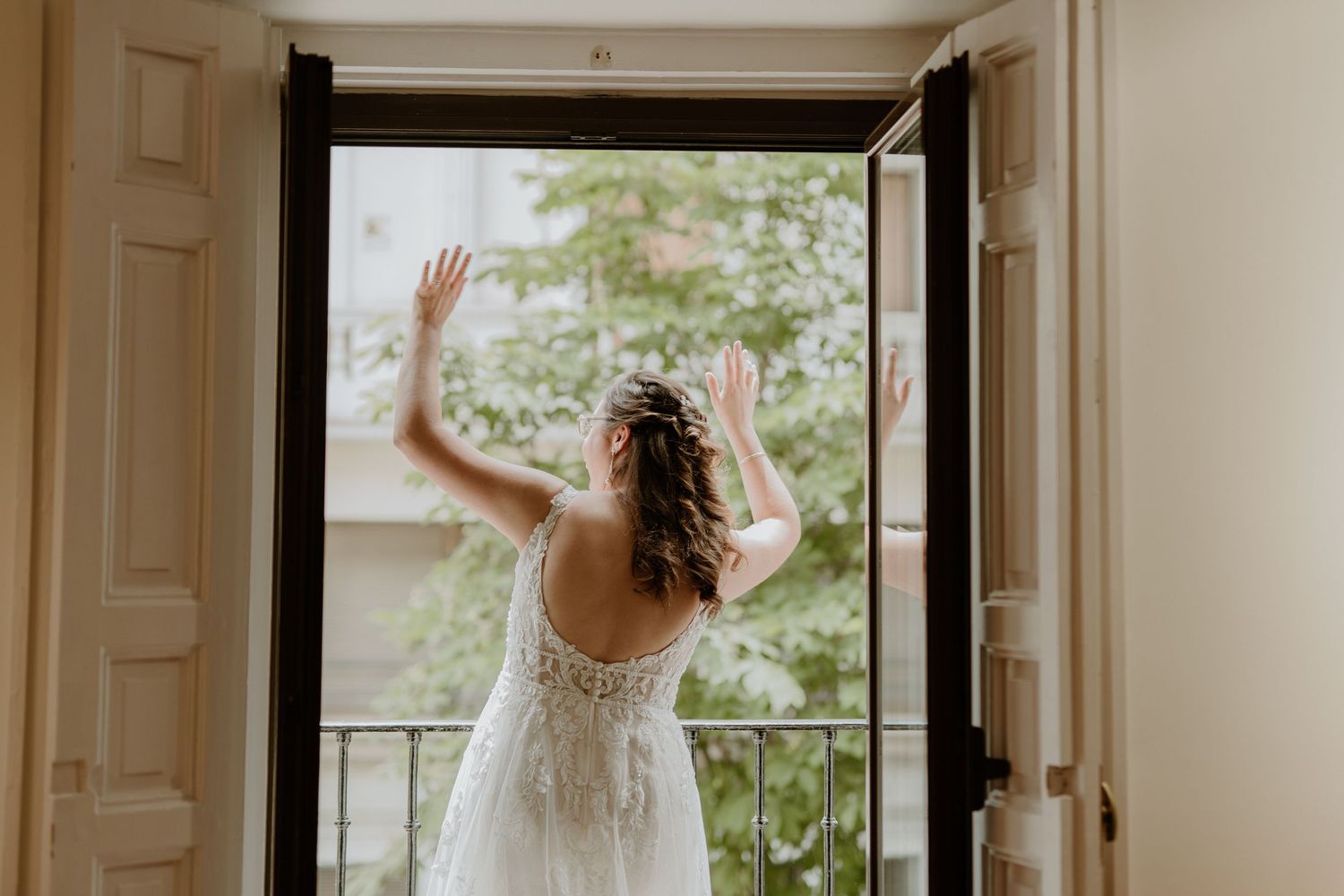 A bride in a delicate lace gown stands at a balcony doorway with her back turned, raising her hand in greeting.