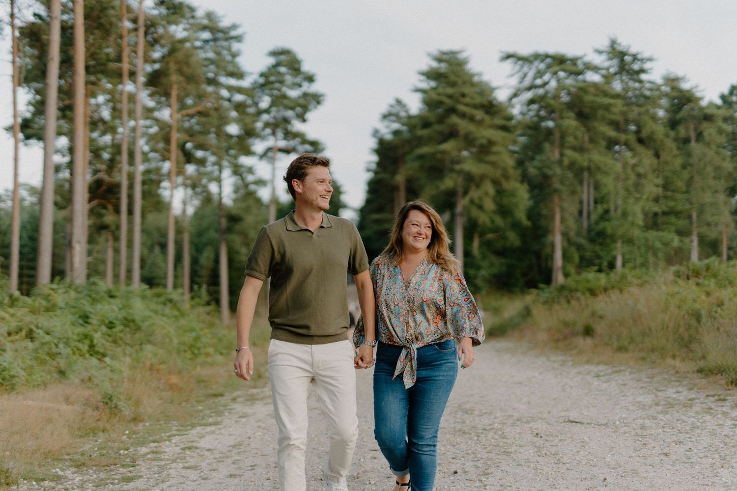 A couple walks together on a forest path surrounded by tall pine trees on a sunny day.