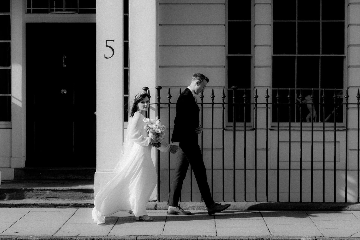 A couple walking past elegant white townhouses with black railings on their wedding day, captured in black and white.