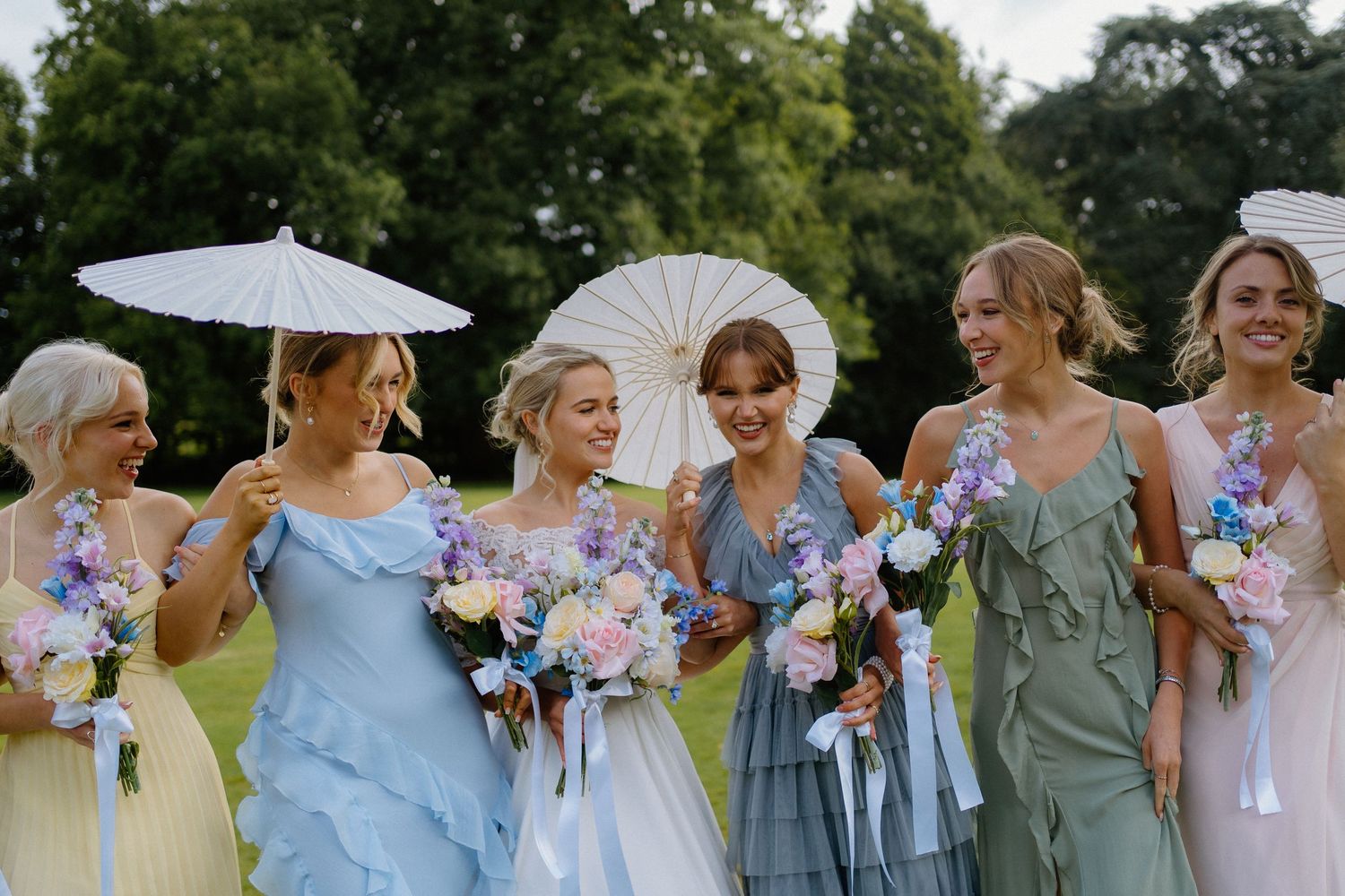 Bridesmaids in pastel blue and green dresses holding floral bouquets and white parasols outdoors with a bride in white.