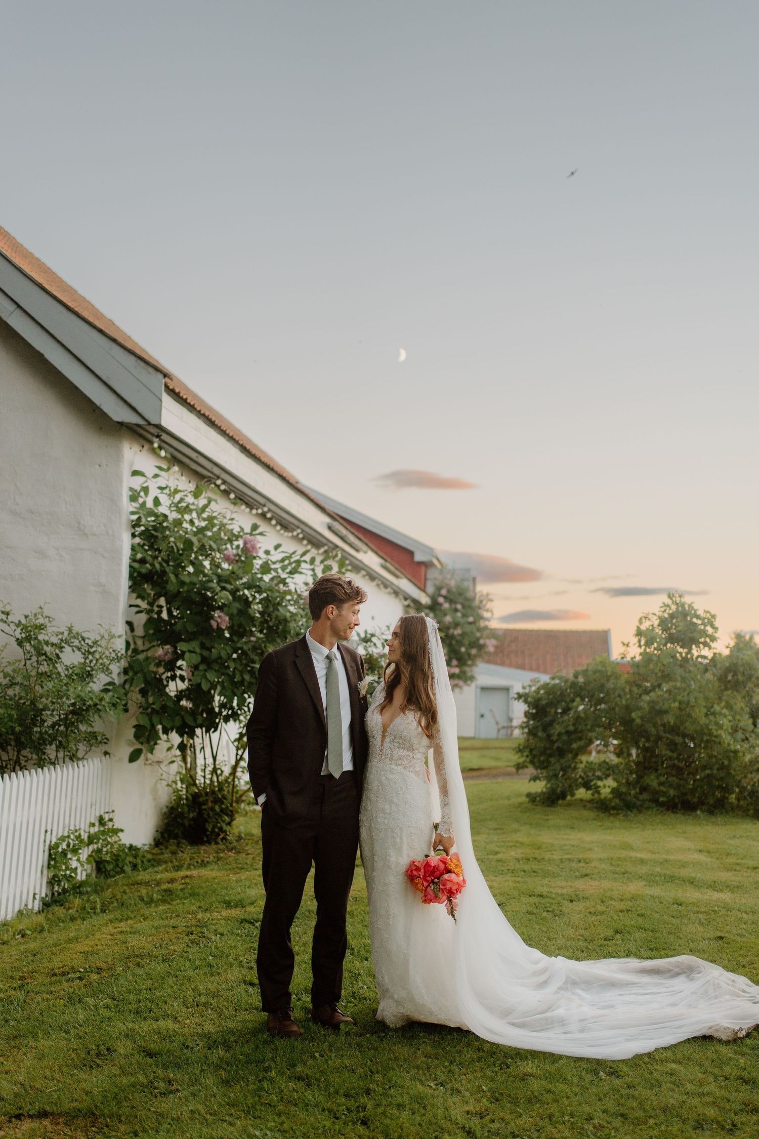 Bride and groom share romantic moment at dusk beside white building, bride holding bright coral bouquet.