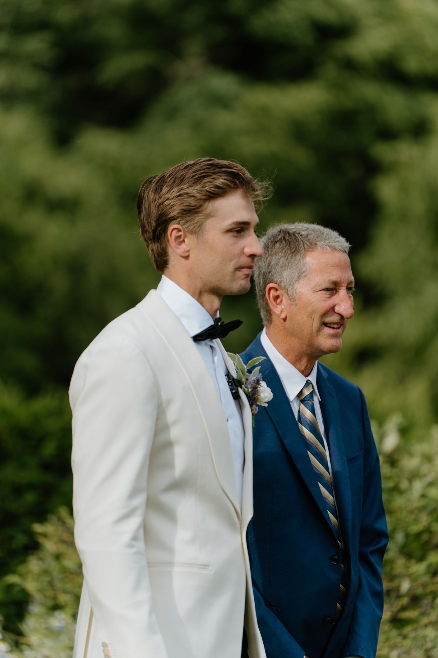 Groom in white tuxedo stands at altar with man in blue suit during outdoor wedding ceremony.
