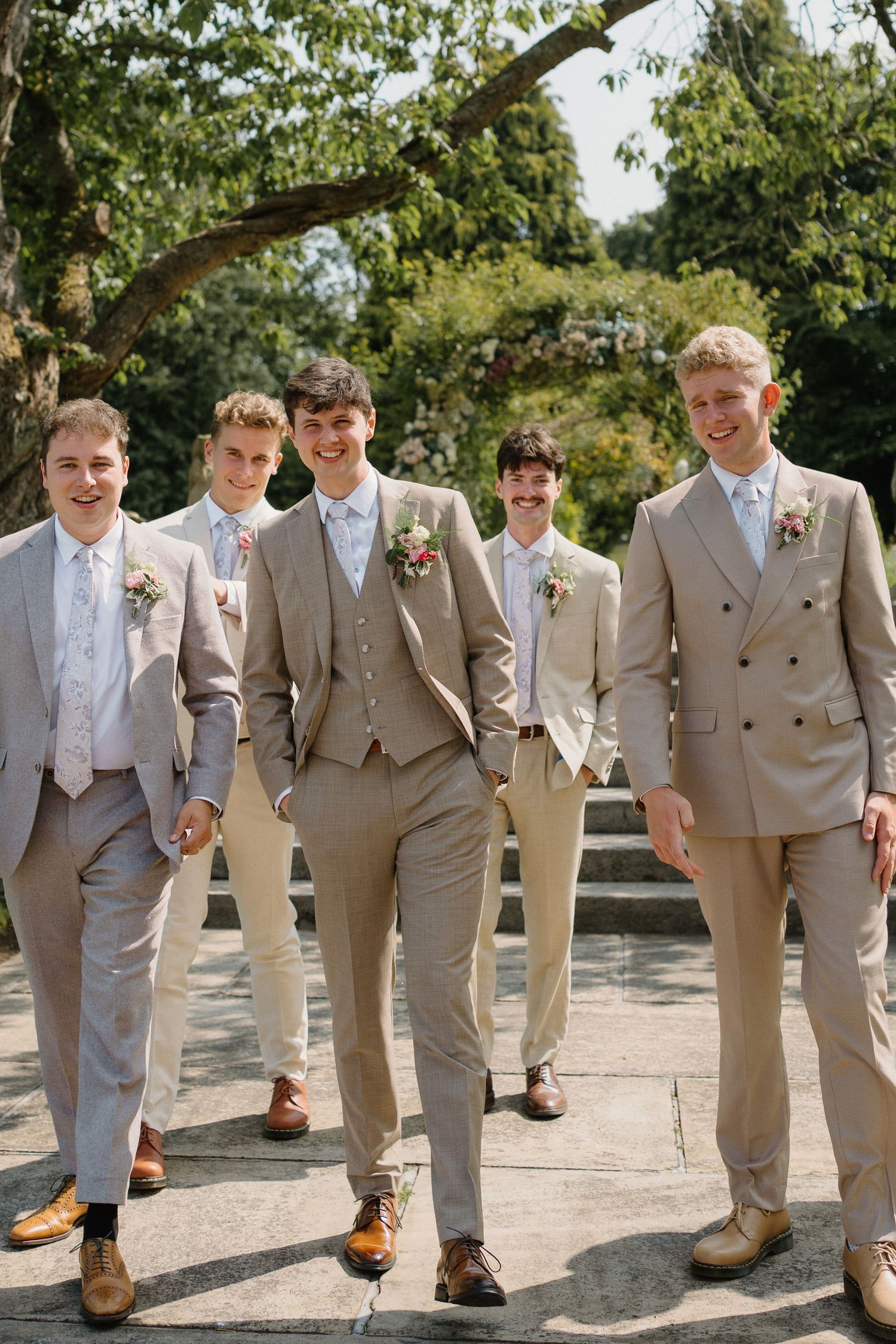 Five groomsmen in matching beige suits with floral boutonnieres walking outdoors on sunny day.