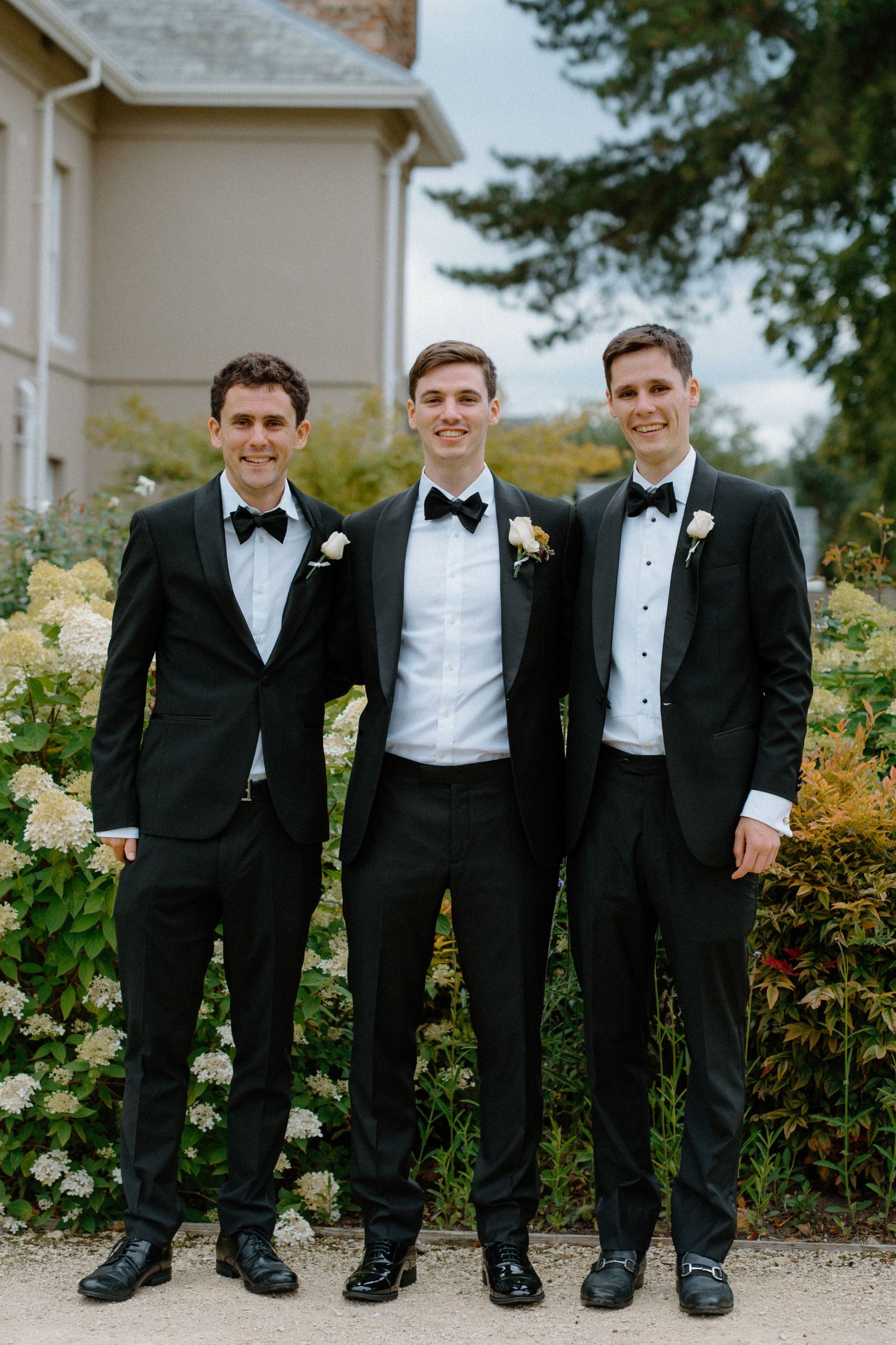 Three groomsmen in black tuxedos with bow ties and boutonnieres posing outdoors at wedding venue.