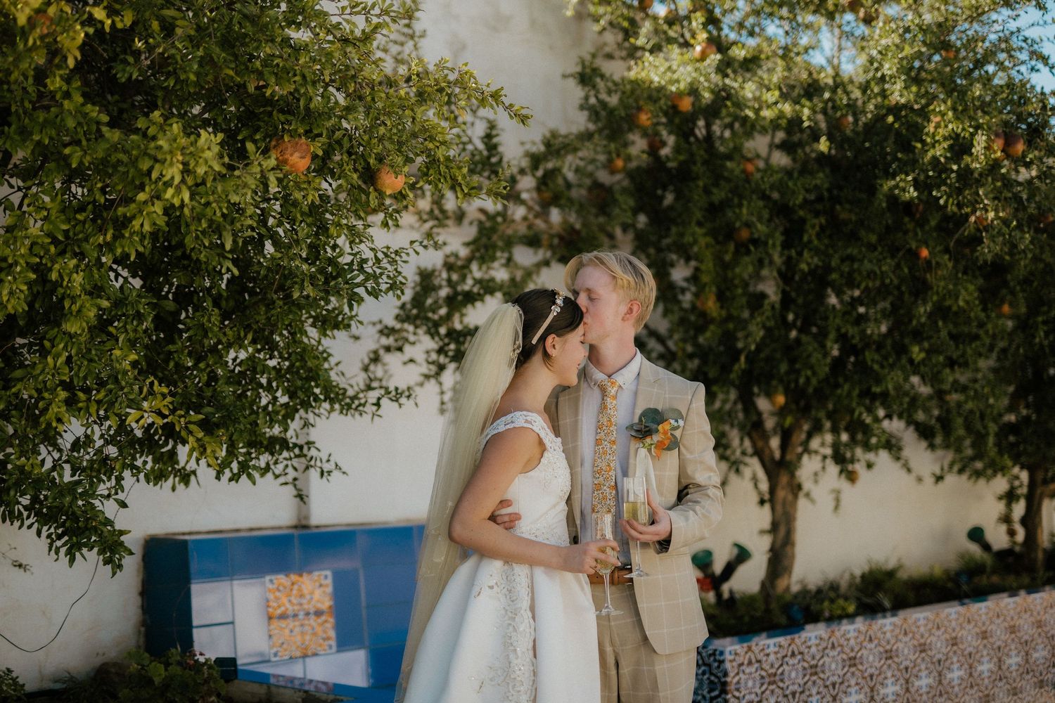 Couple shares tender moment under orange tree at outdoor wedding venue.