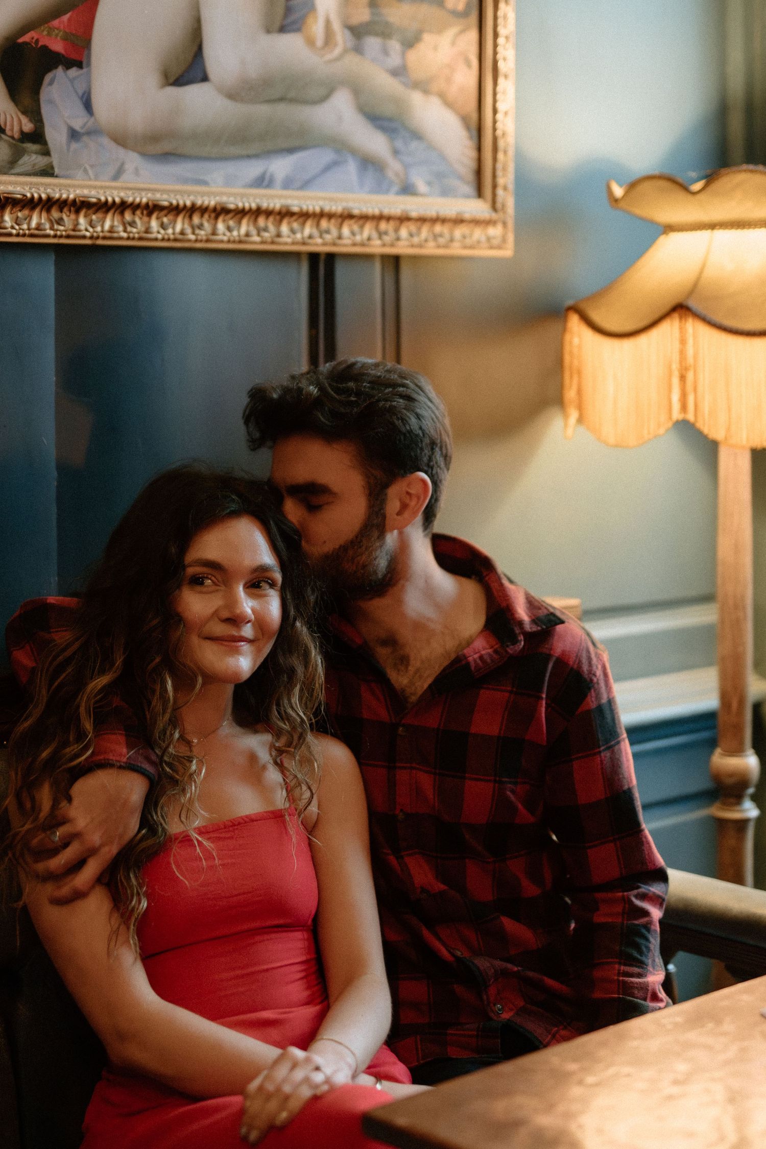 Couple sharing a tender moment on a velvet chair beside a vintage lamp in a moody interior.
