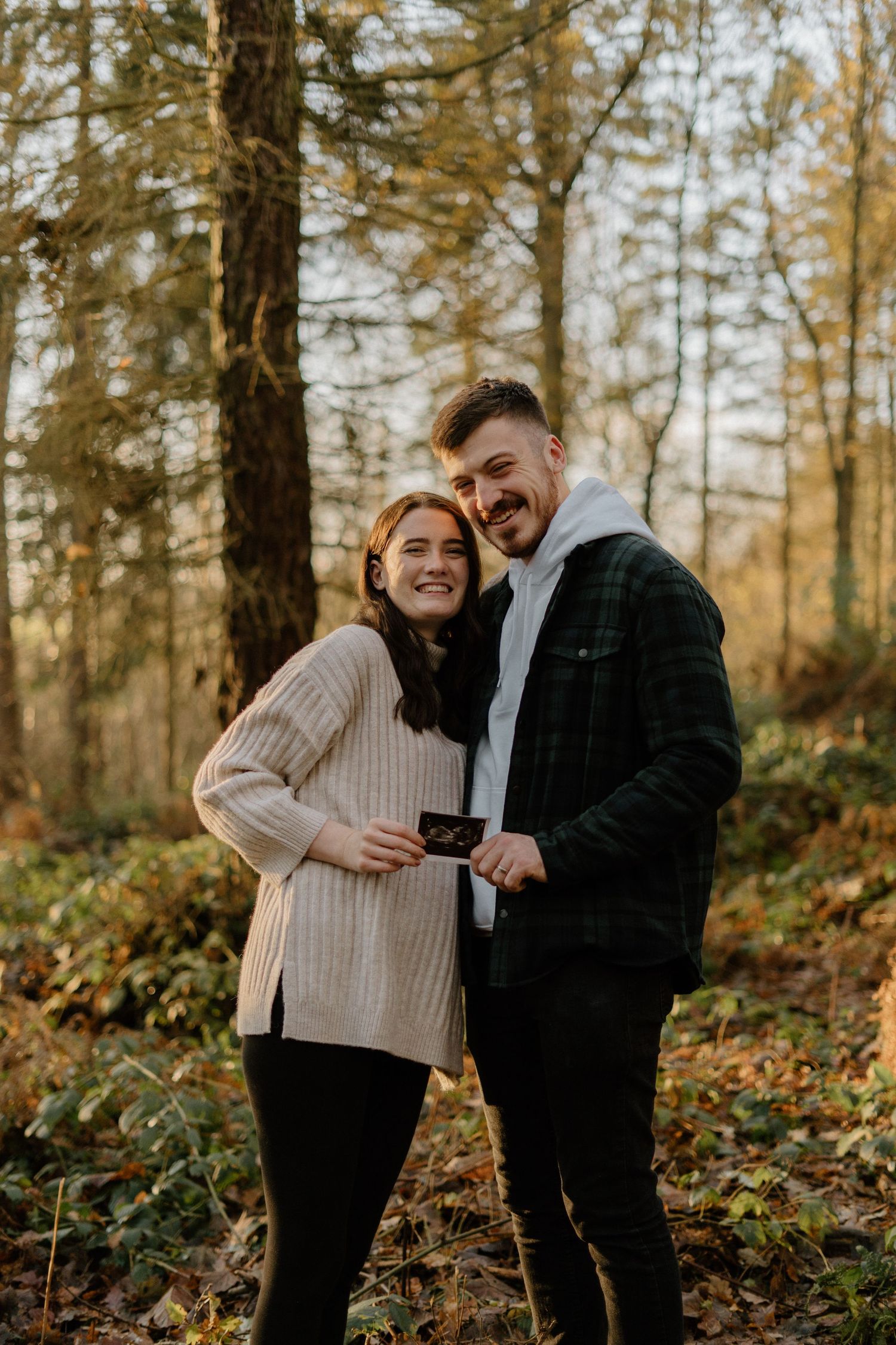 Smiling couple holding ultrasound scan surrounded by golden autumn forest foliage.