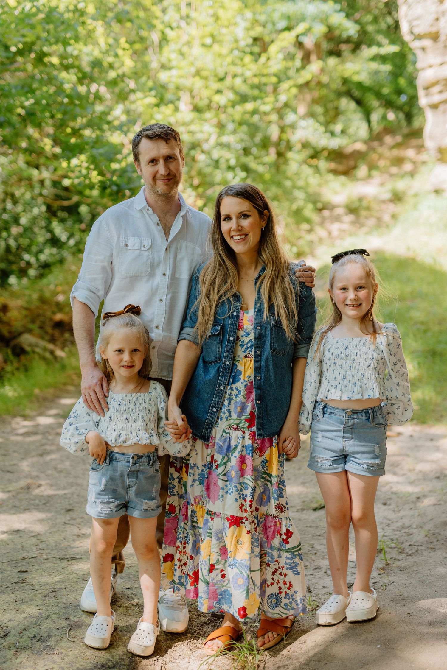 A family of four smiles together on a sunny woodland path surrounded by green trees.