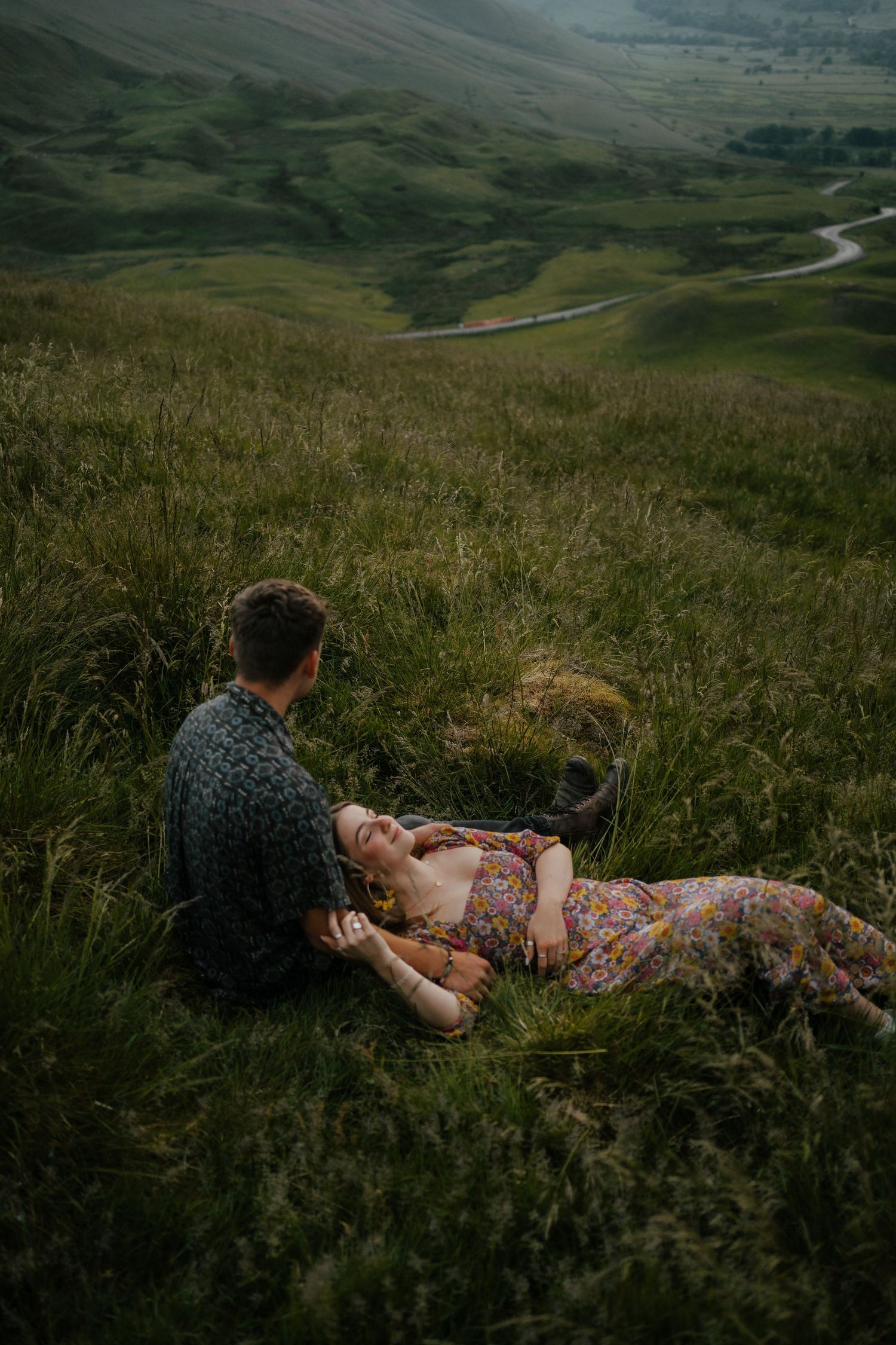 Romantic couple relaxing in green hillside grass with a winding road and valley below.