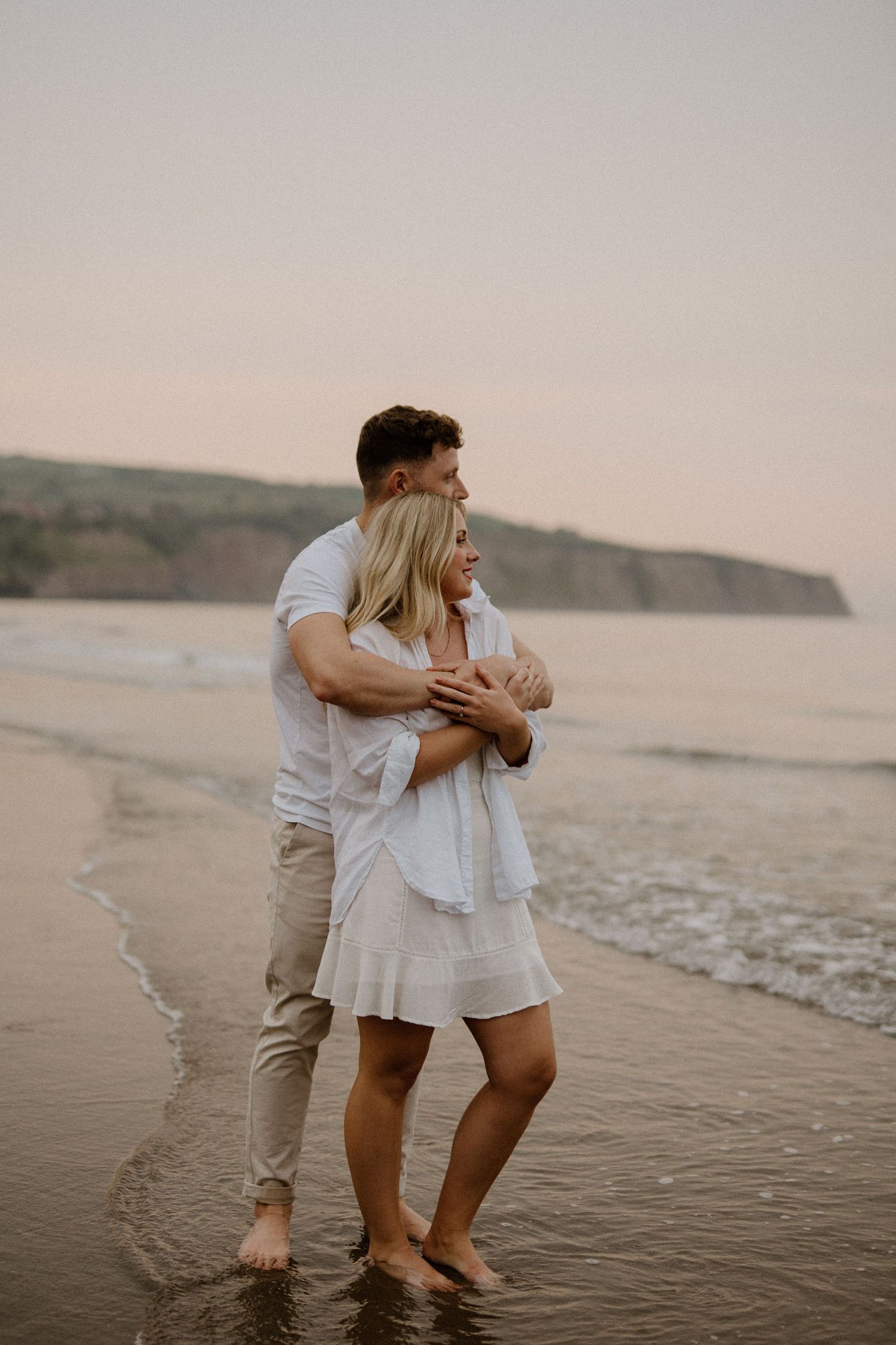 Couple embracing barefoot on a sandy beach at sunset with coastal cliffs in the background.