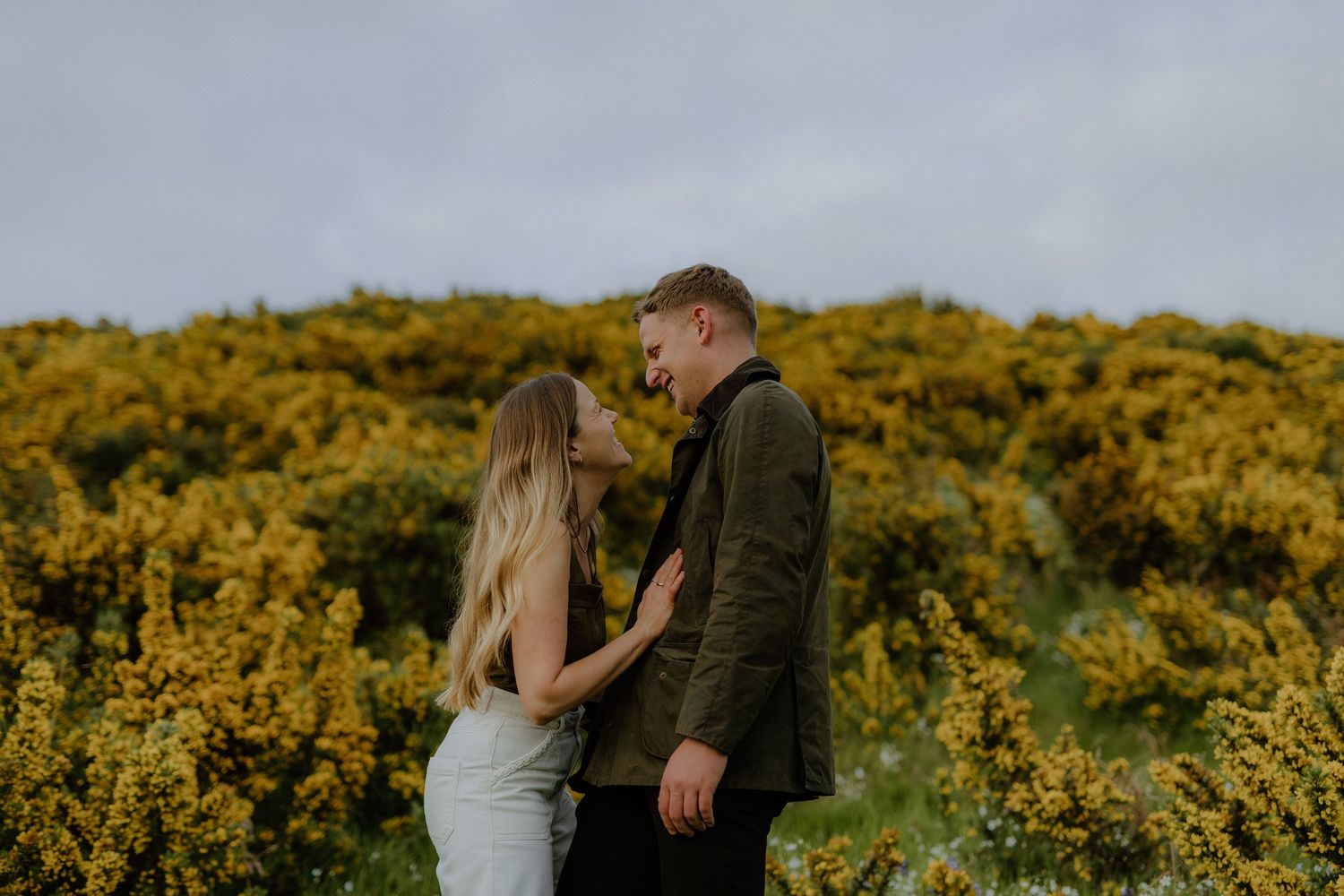 Couple smiling at each other in front of vibrant yellow gorse bushes on a hillside.