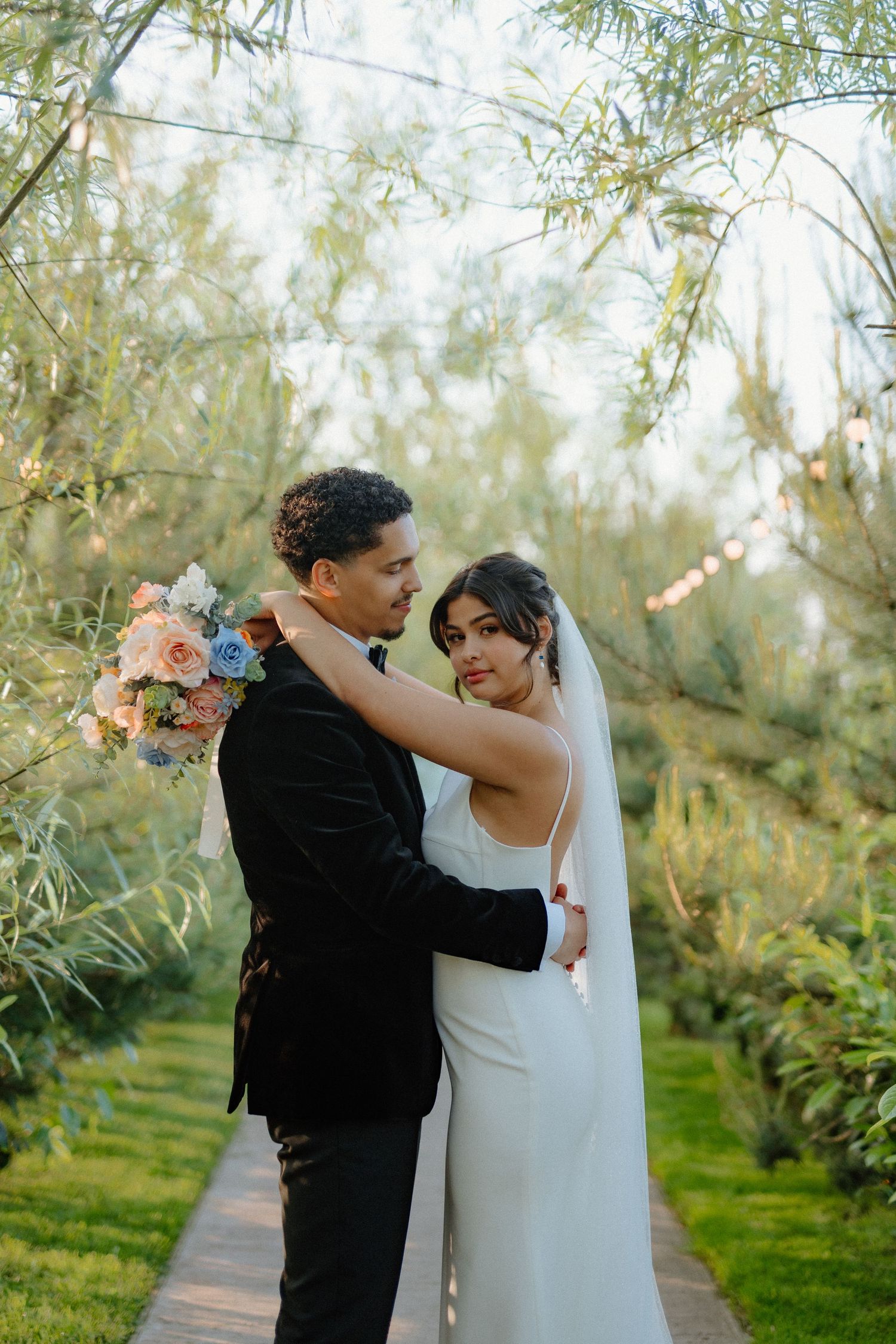 Bride in white gown with veil holds colorful bouquet while embracing groom in black tuxedo in lush garden.