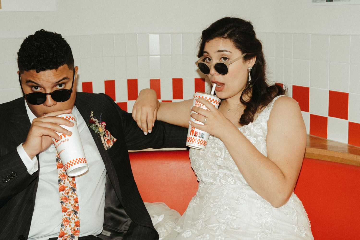 Bride and groom wearing sunglasses sipping drinks together in a red and white fast food restaurant.