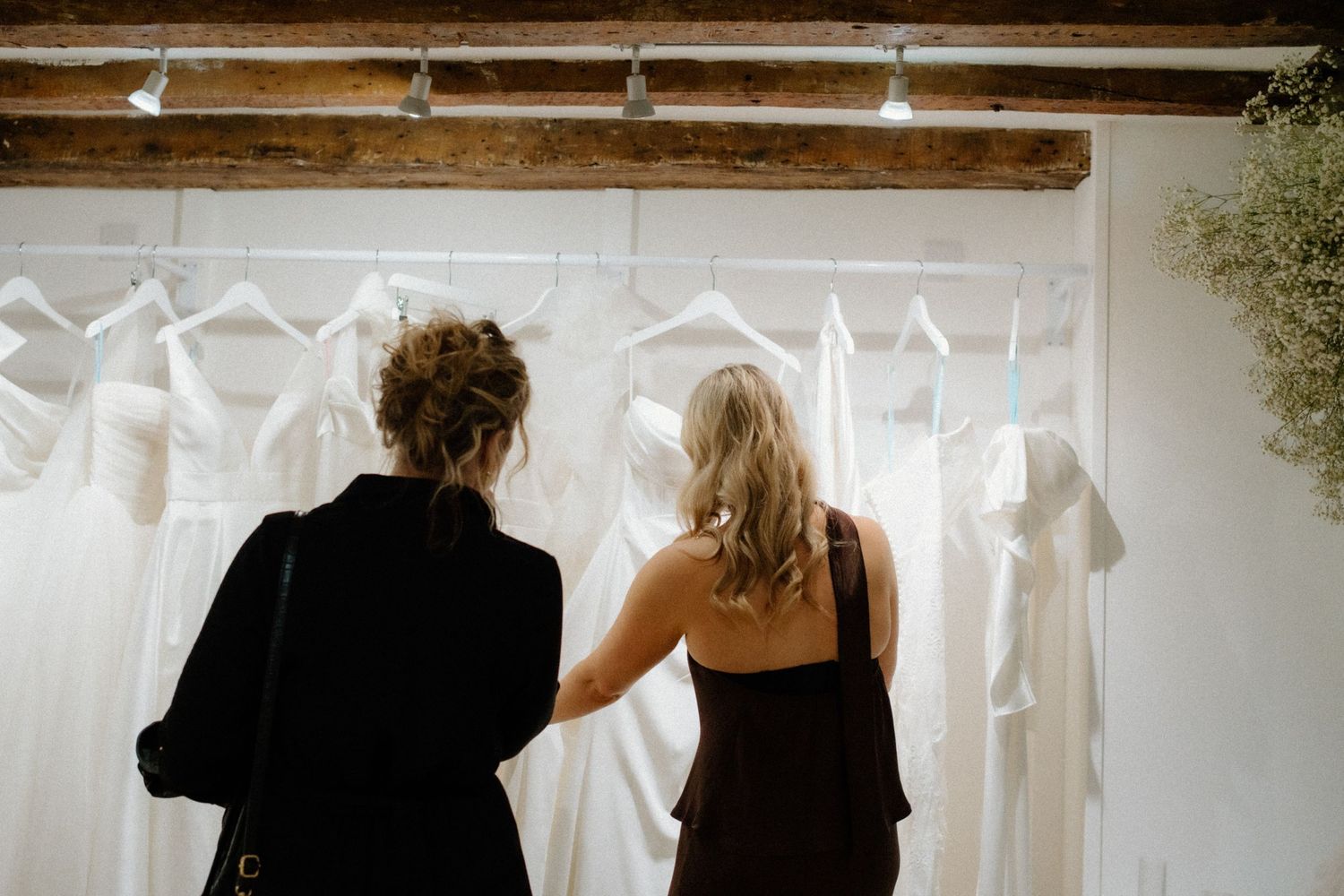 Two people in dark clothing look at white dresses hanging on a rack against a white wall beneath rustic wooden beams.