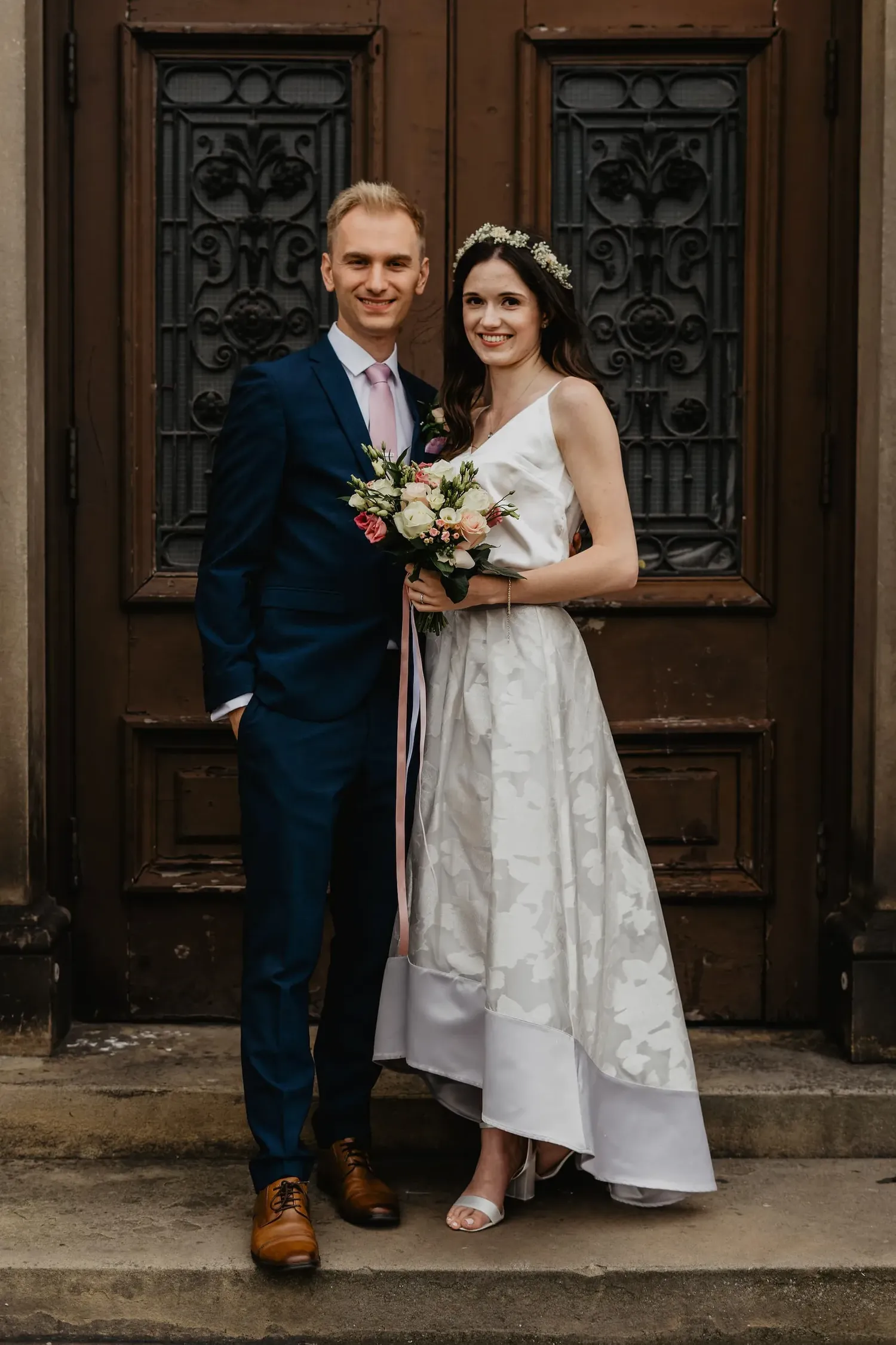 A couple poses for a wedding photo in front of vintage wooden doors wearing navy suit and tea-length dress.