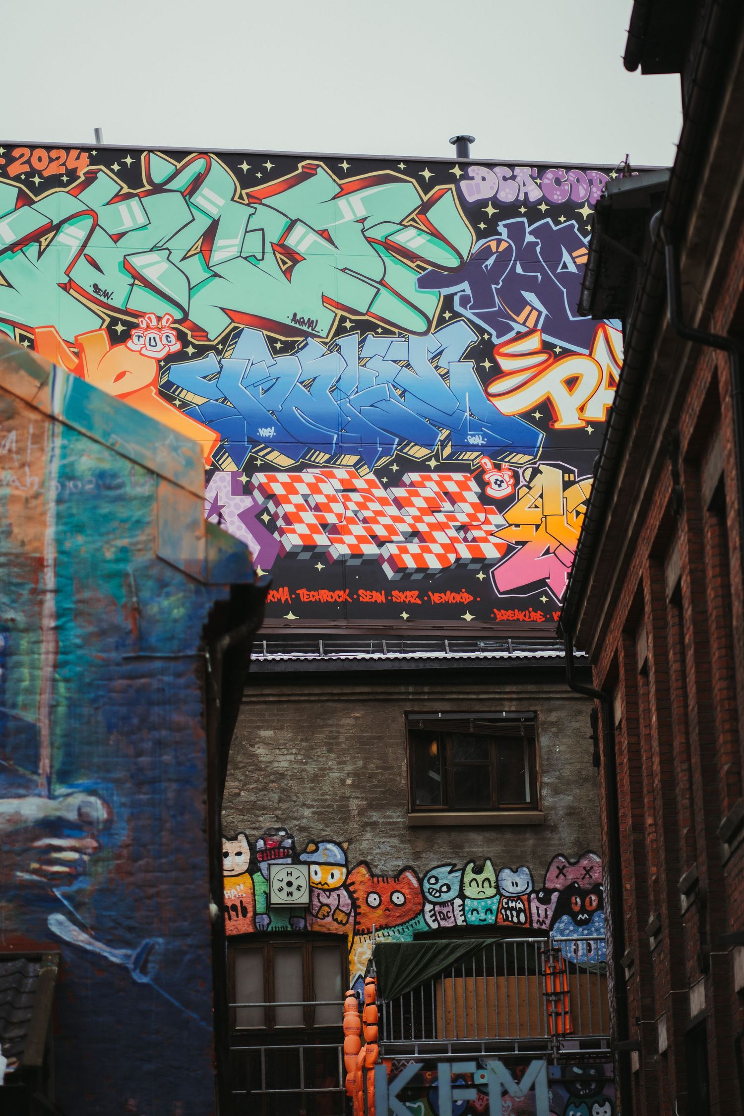 Colorful street art graffiti adorns the walls and overhangs of an urban alleyway with brick buildings.