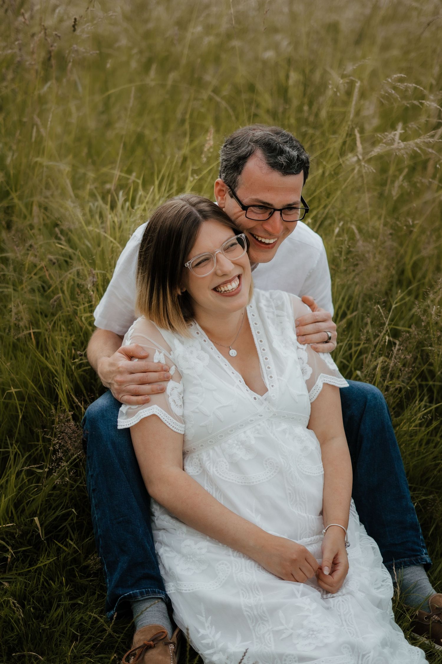 A happy couple sits in tall grass, the man embracing a smiling pregnant woman in white.