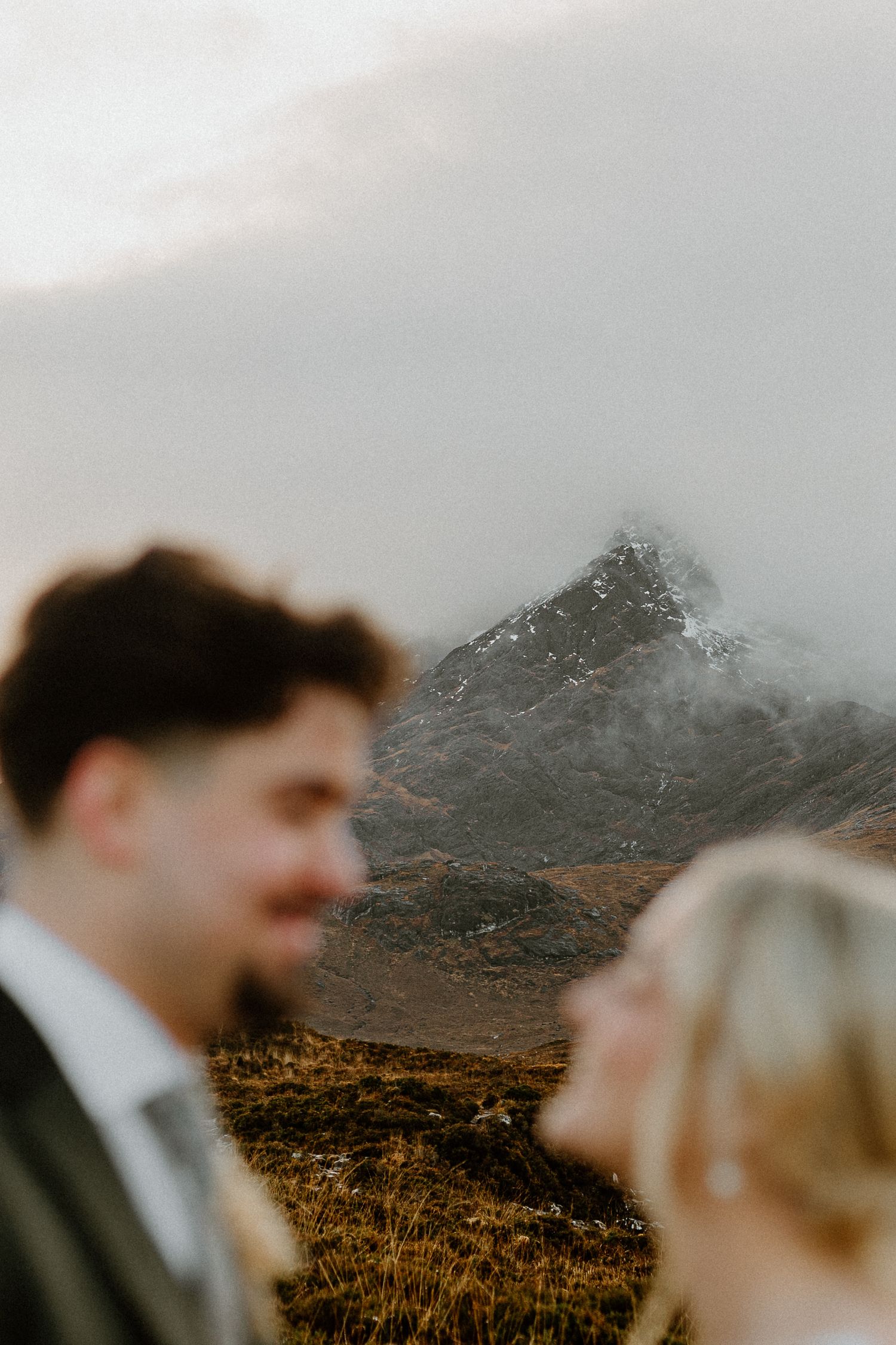 Artistic blurred foreground of bride and groom with dramatic misty Scottish mountain peak in sharp focus behind them.