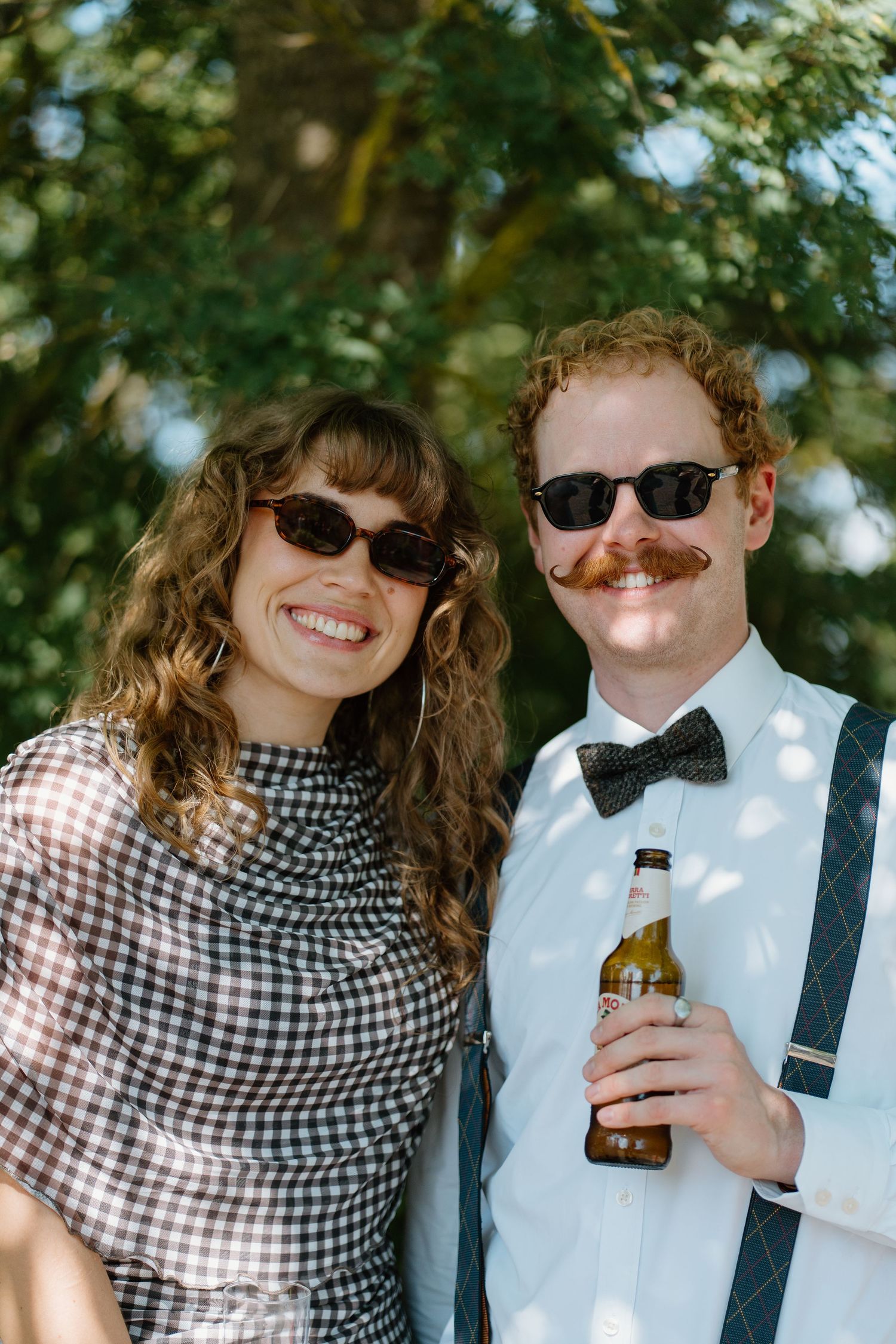 Smiling wedding guests wearing sunglasses and bow tie holding beer bottle at outdoor reception.