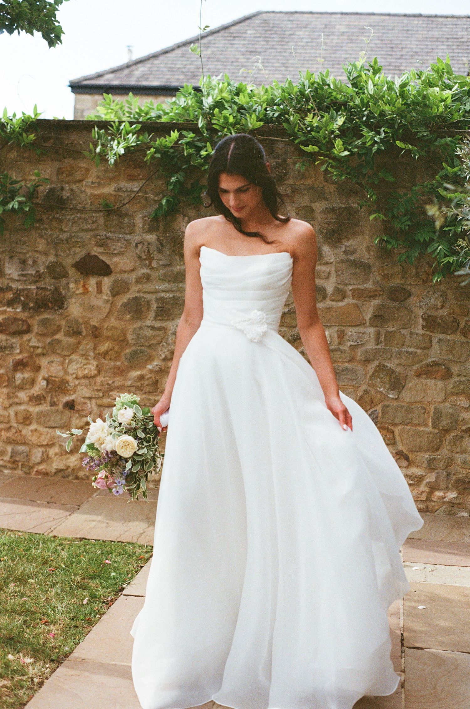 Bride in strapless white ball gown holding bouquet against rustic stone wall with green ivy.