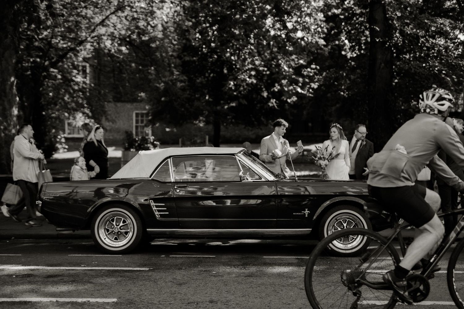 Black and white photo of a wedding party with a classic Ford Mustang on a tree-lined street.