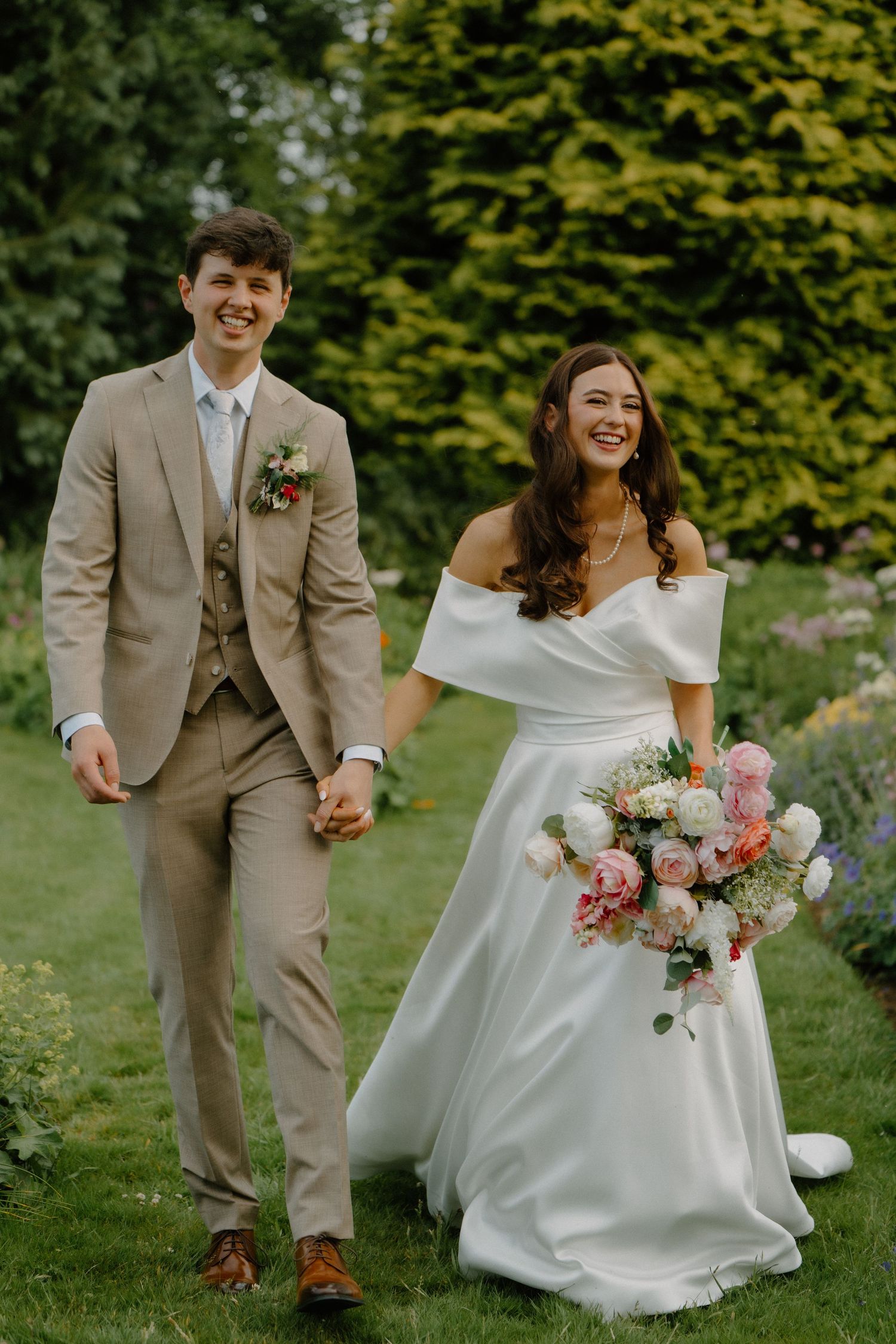 Smiling bride and groom walking hand in hand through a lush garden, bride holding a colorful bouquet in a white off-shoulder gown.