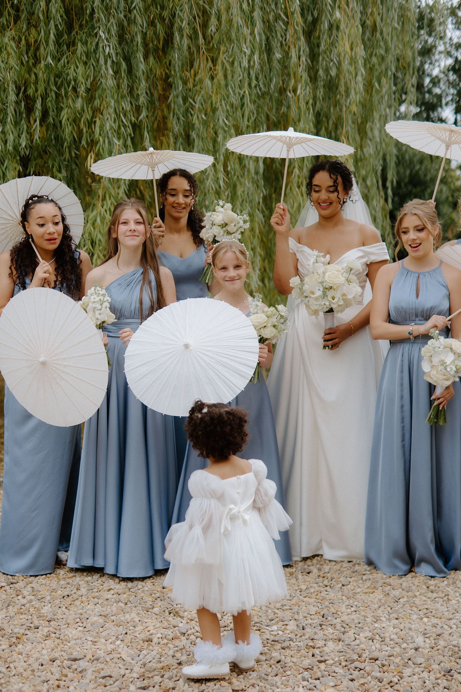 Bride poses with bridesmaids in blue gowns holding white parasols with flower girl in foreground.