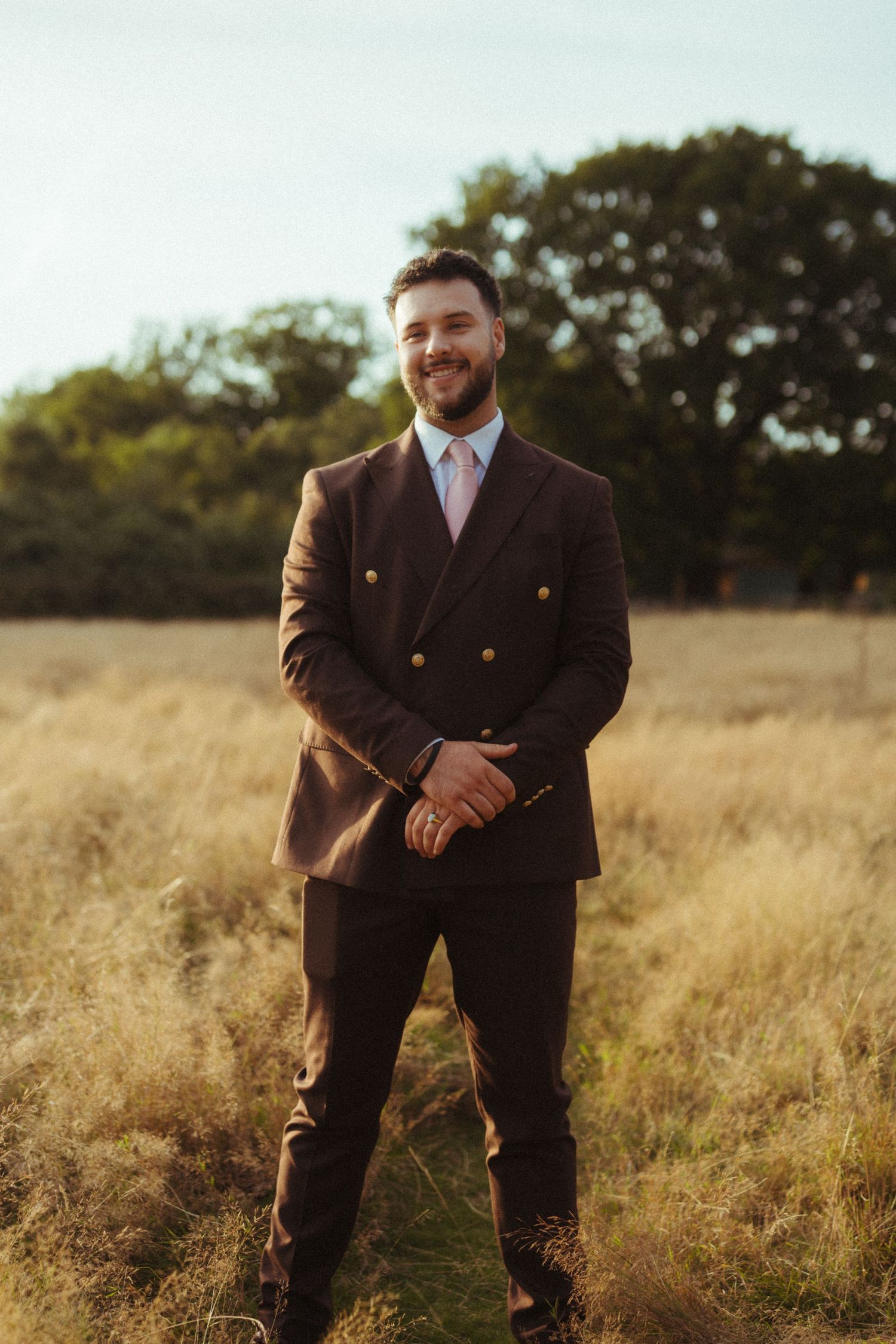 Handsome groom in brown double-breasted suit with pink tie stands smiling in golden meadow at sunset.