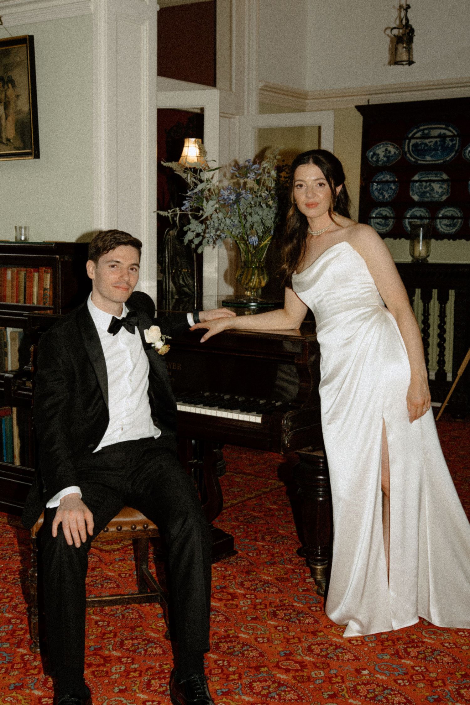 Bride in elegant white satin gown and groom in tuxedo posing beside a grand piano.