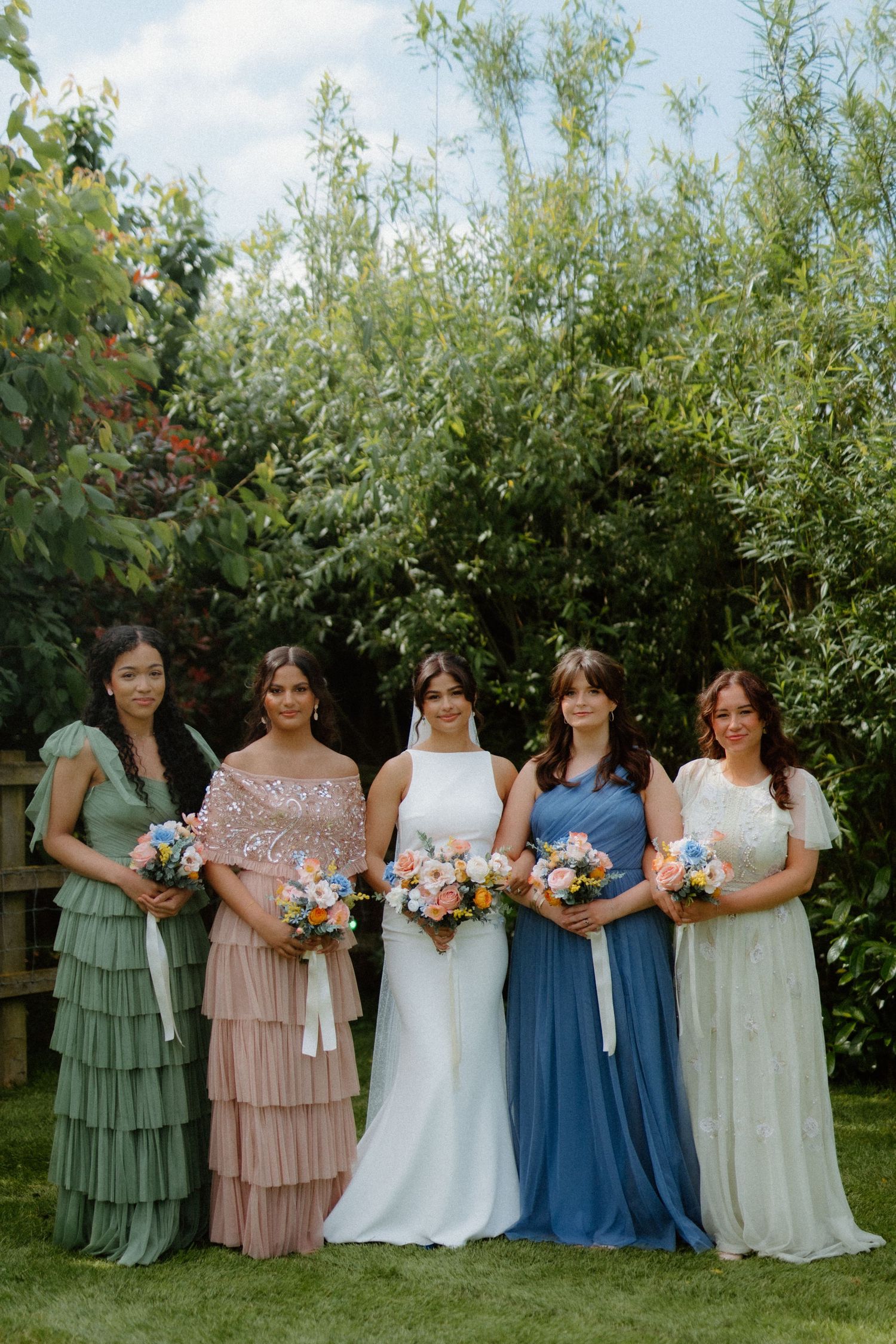 Bride with four bridesmaids in mismatched pastel gowns holding colorful bouquets in a garden.