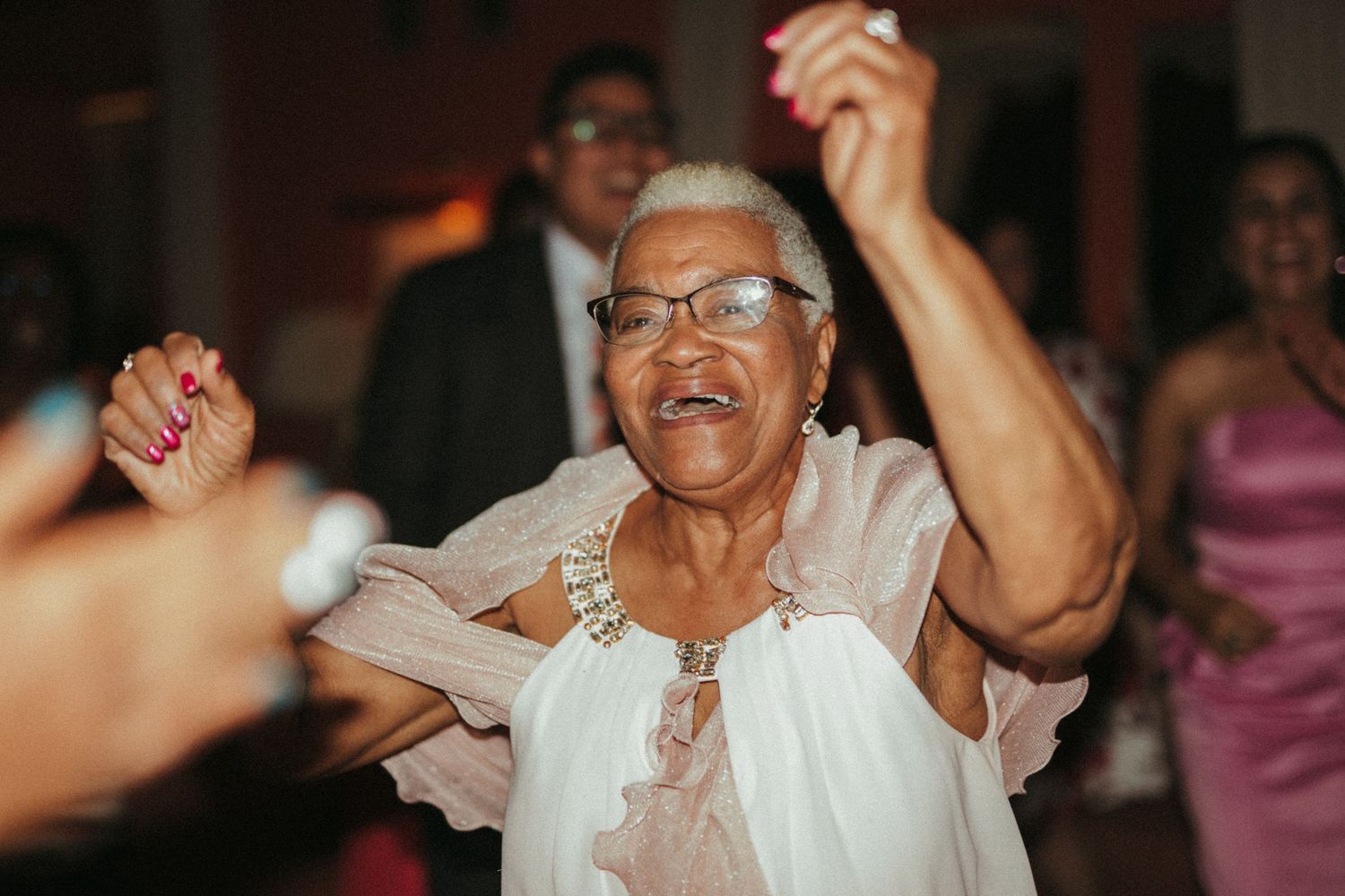 Elderly woman in white dress dancing joyfully with arms raised at a festive wedding reception.