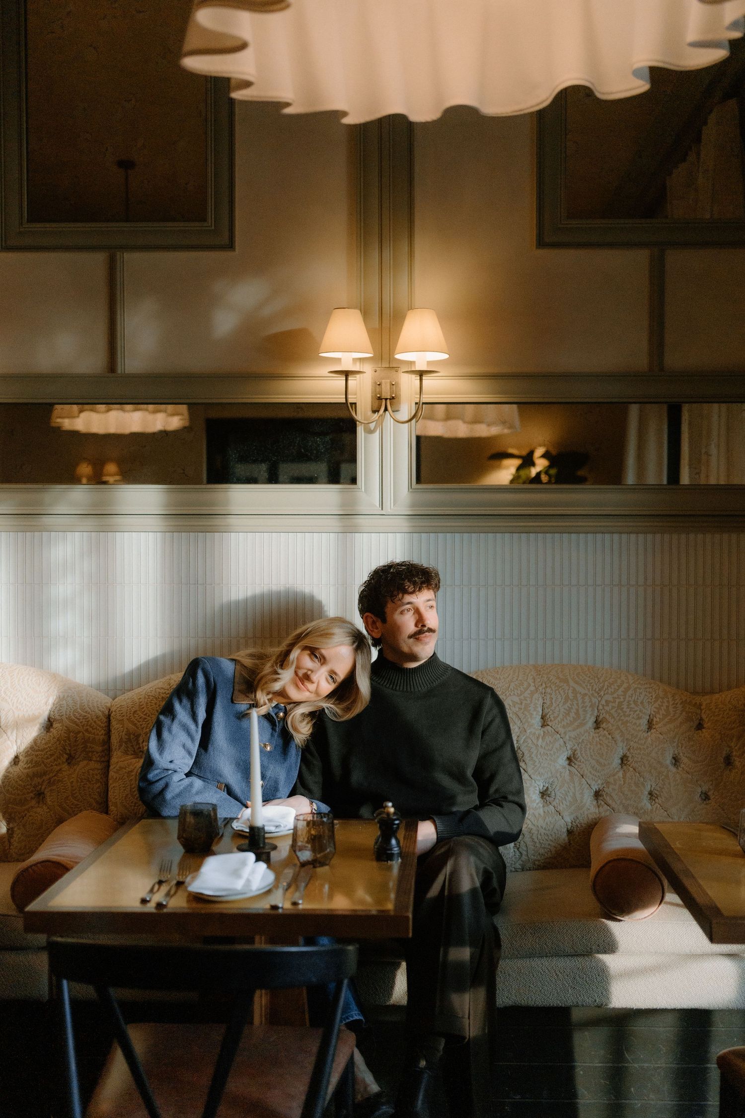Stylish couple sits together on tufted sofa in warmly lit vintage restaurant booth setting.