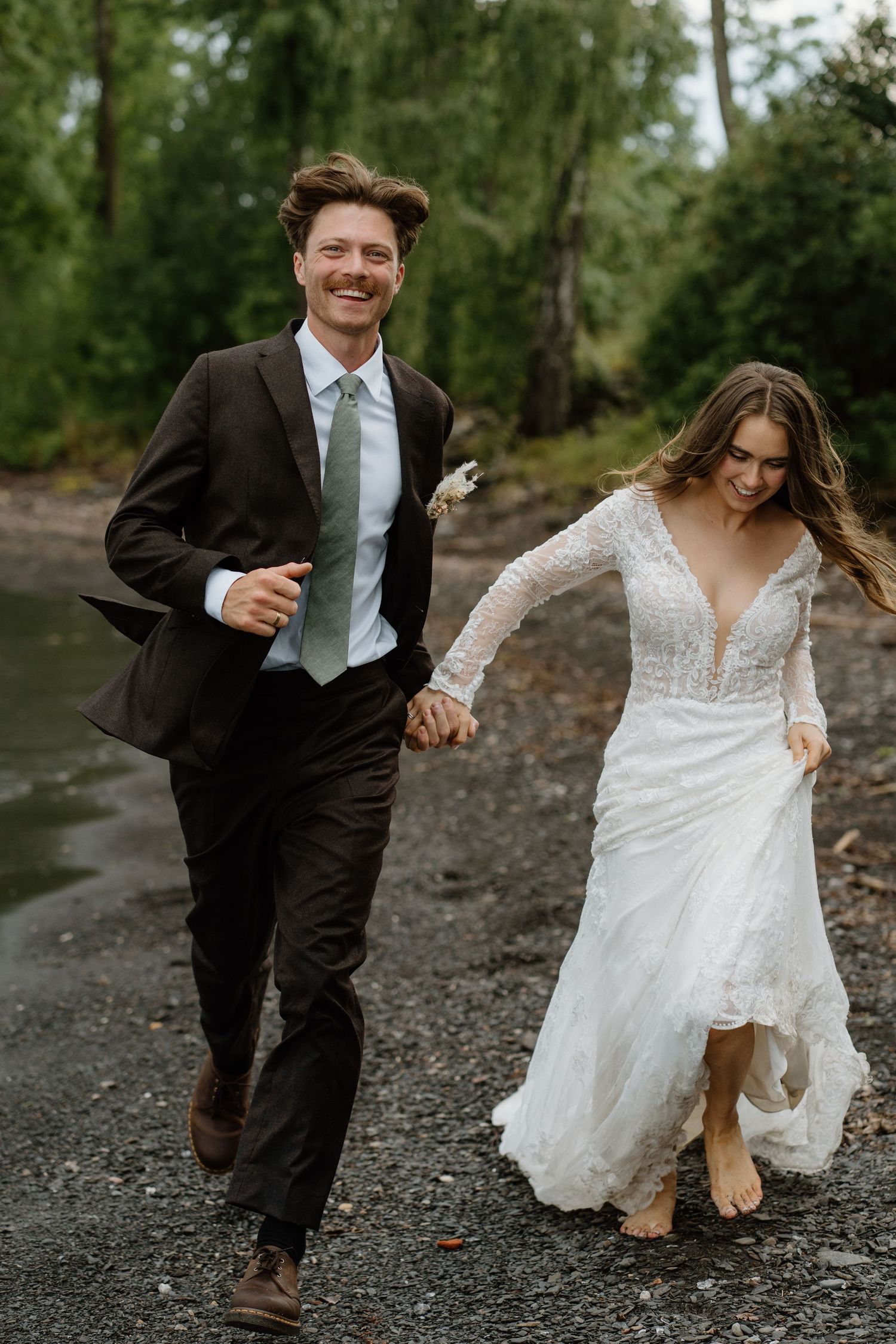 Joyful bride and groom run hand-in-hand barefoot along scenic rocky beach with lush forest.