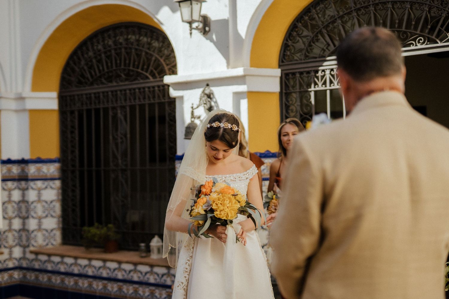 Bride holding yellow bouquet in white gown and veil at colorful Spanish courtyard entrance.