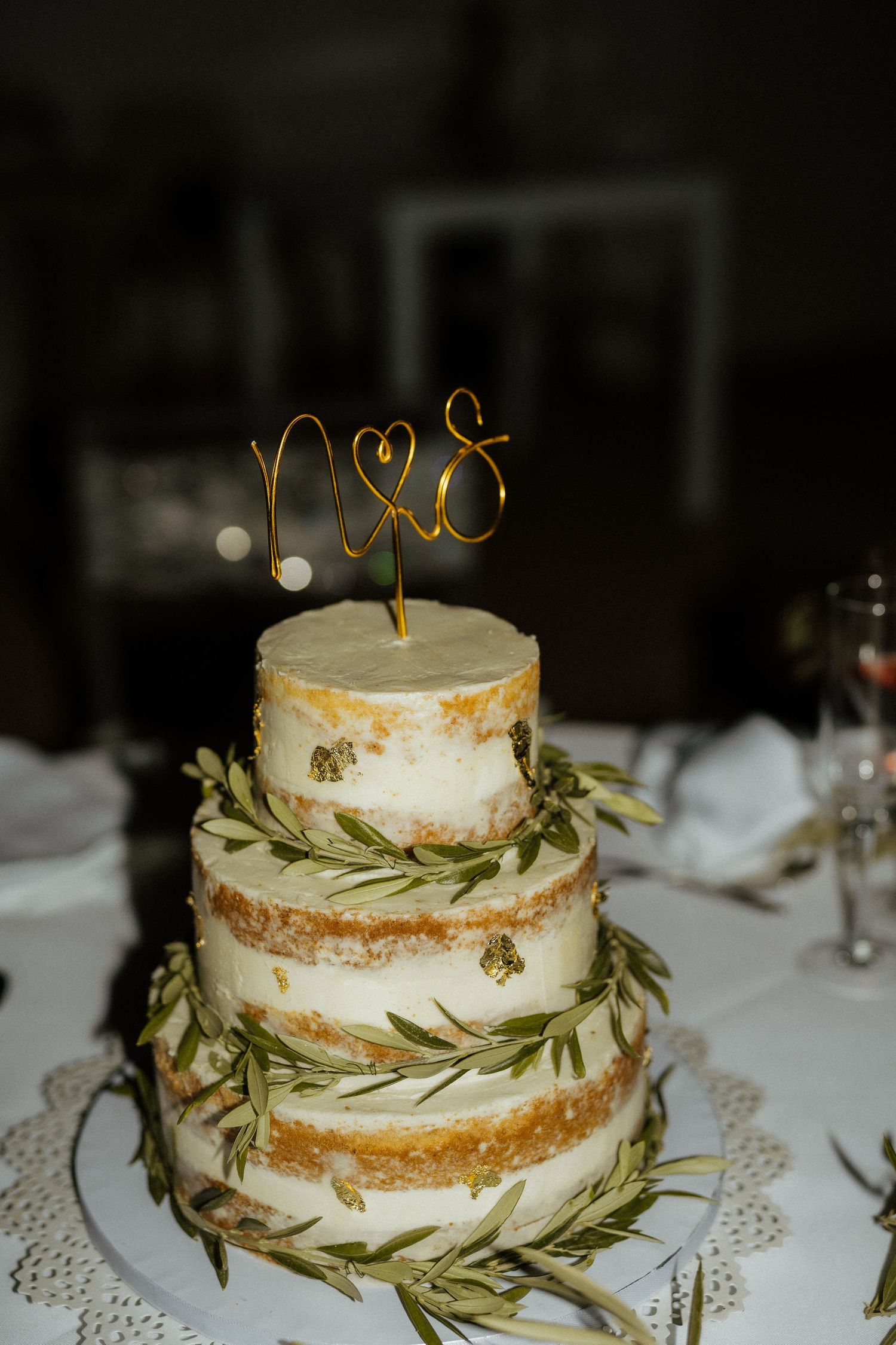 Three-tier naked wedding cake with rosemary garlands and gold wire Mrs topper on white table.