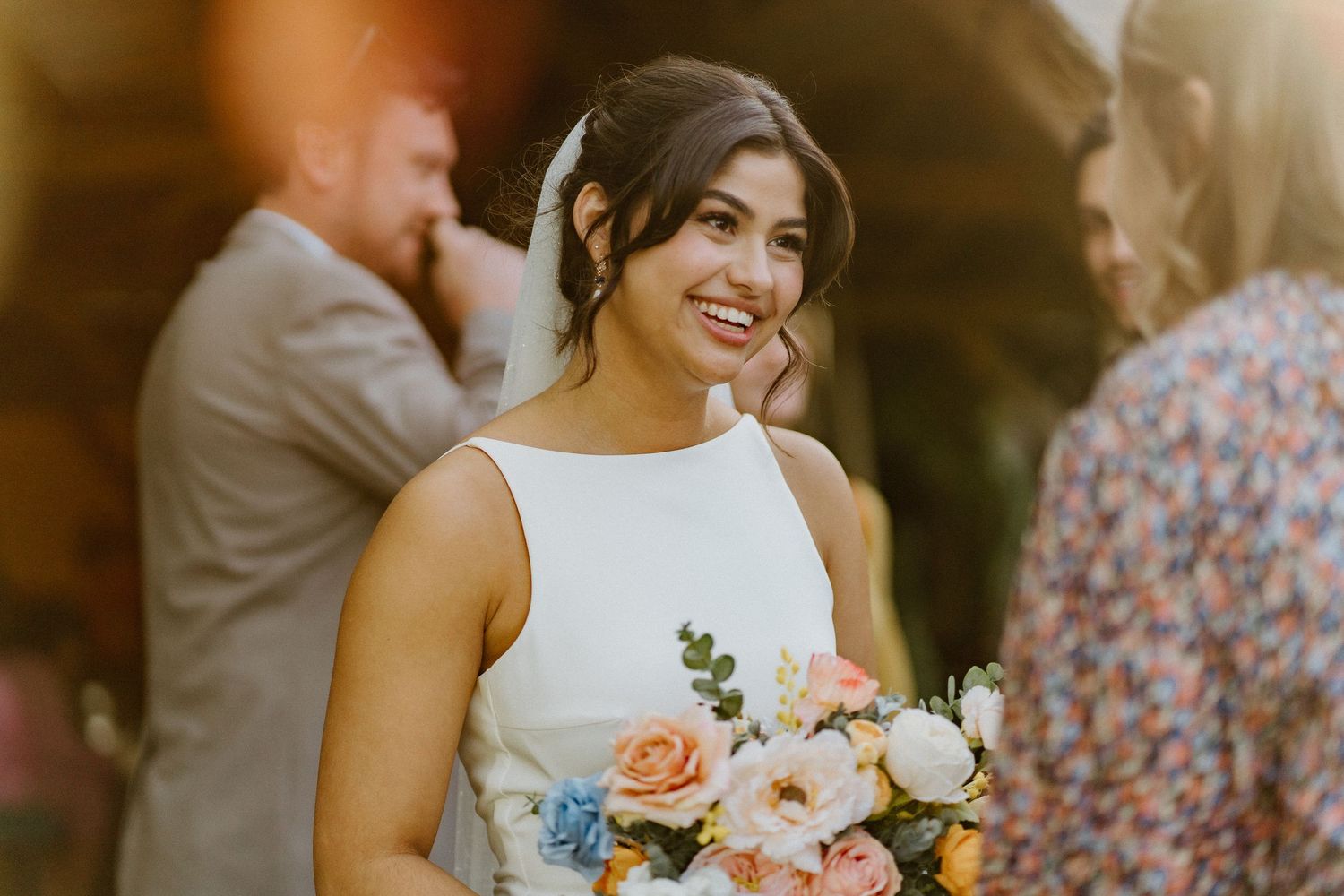 Glowing bride holding pastel floral bouquet and smiling warmly during outdoor wedding reception.