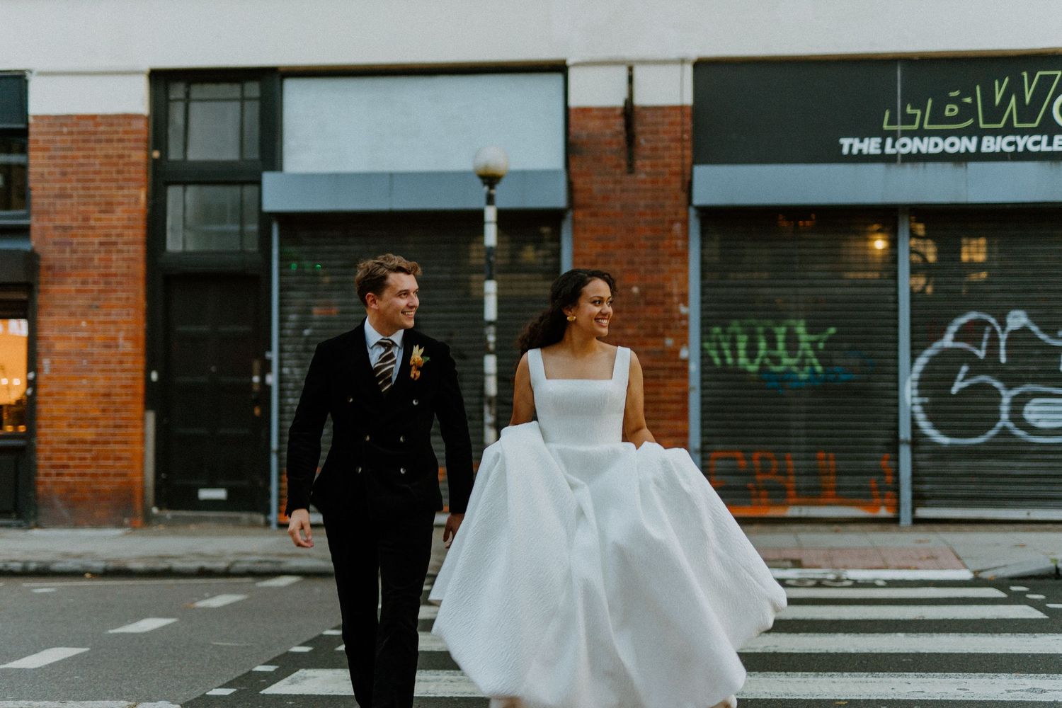 A bride and groom walk hand in hand across a zebra crossing in front of a London bicycle shop.