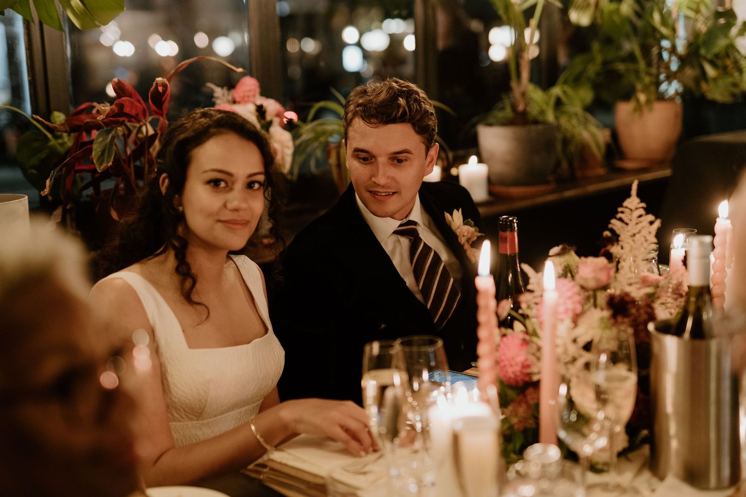 Bride and groom smiling at candlelit table surrounded by floral arrangements at reception.