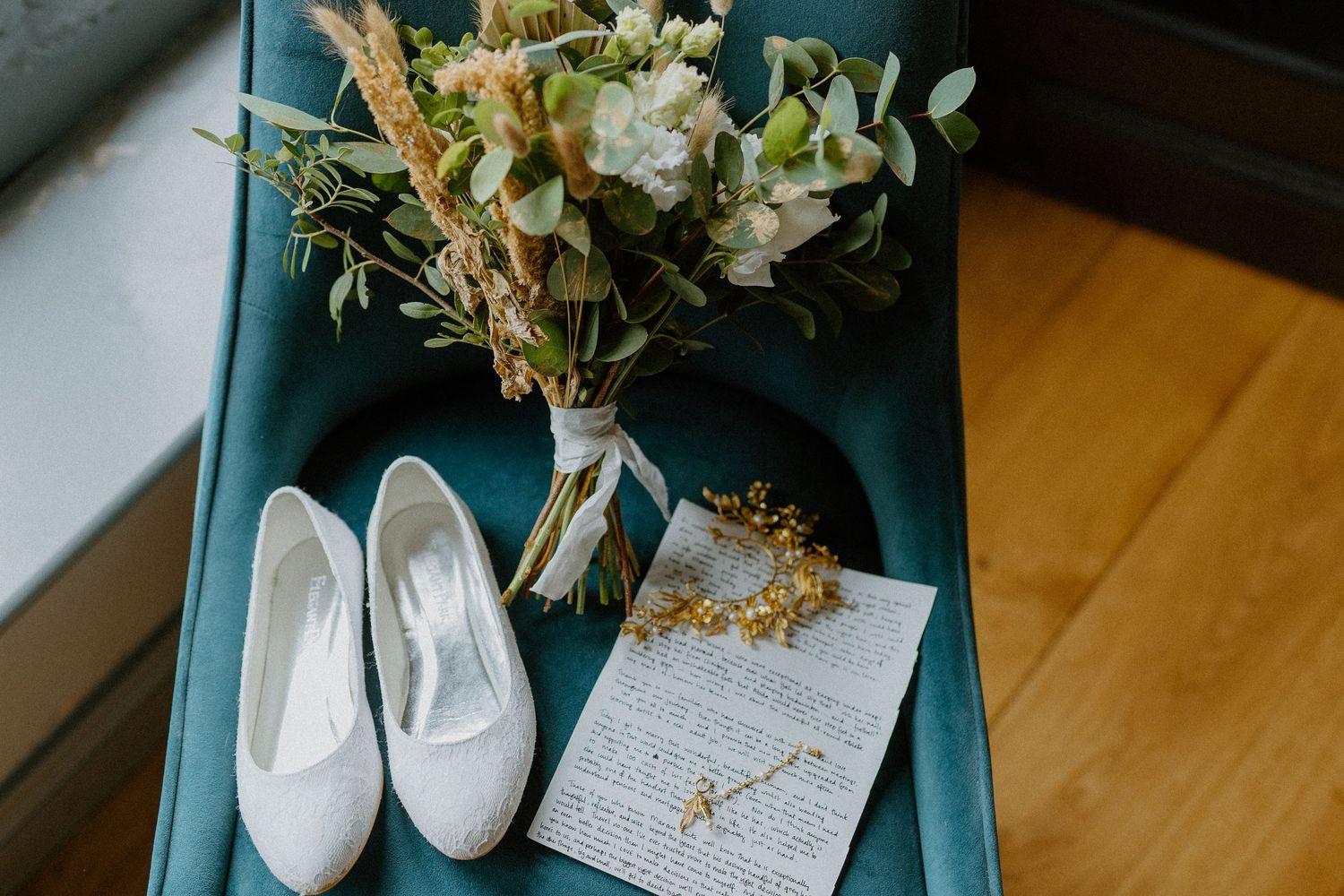 Bridal flat shoes, wildflower bouquet, and handwritten vows arranged on a teal velvet chair.