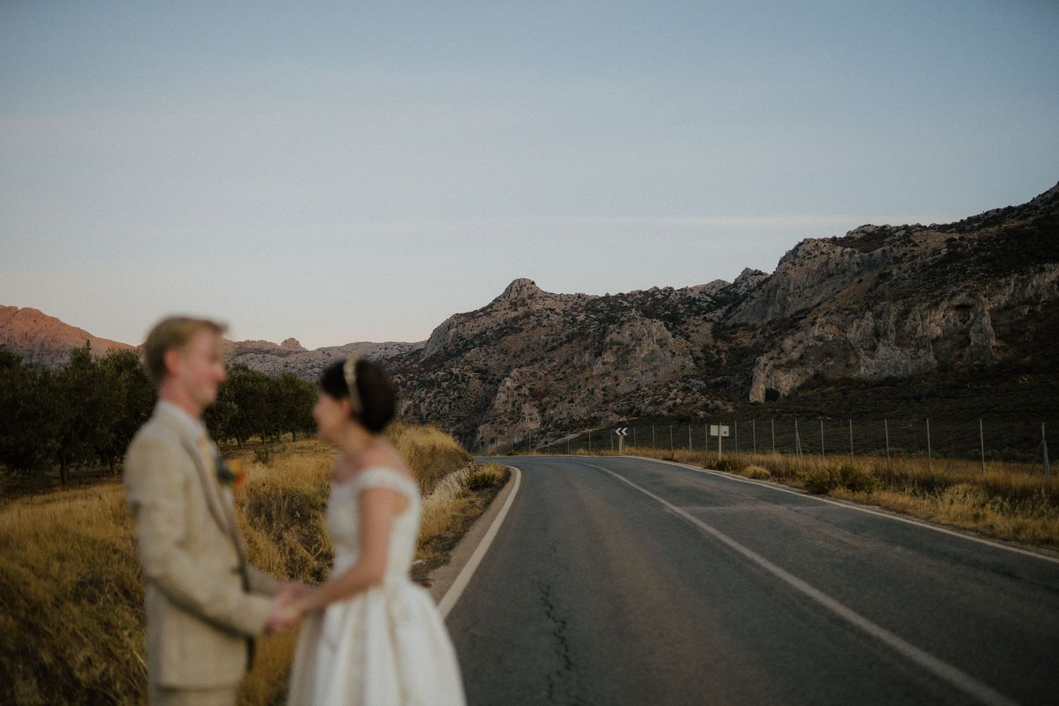 Bride and groom hold hands on a quiet road with dramatic rocky mountains at dusk.