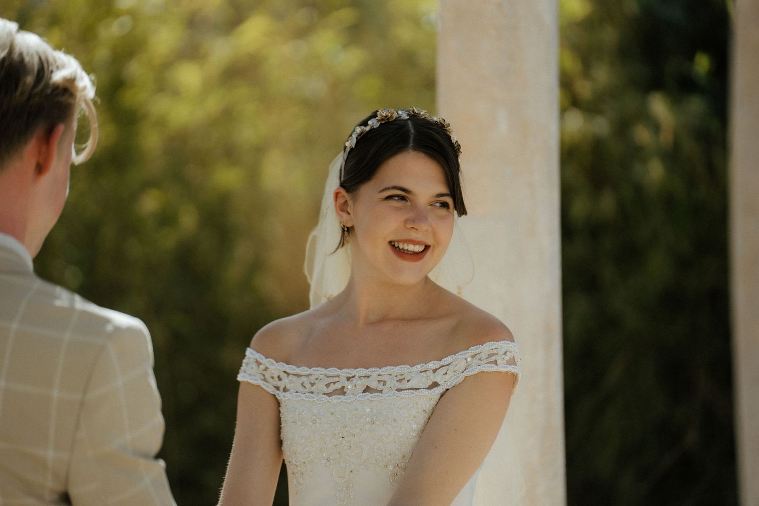 Radiant bride in off-shoulder lace wedding dress smiling joyfully during outdoor wedding ceremony.