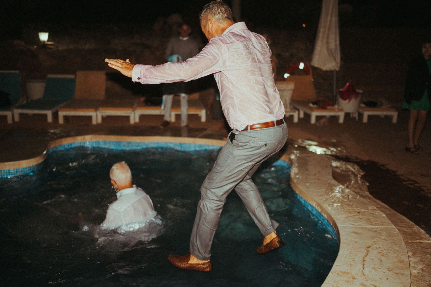 A man in dress clothes stepping into a pool while another person splashes in the water at a wedding.