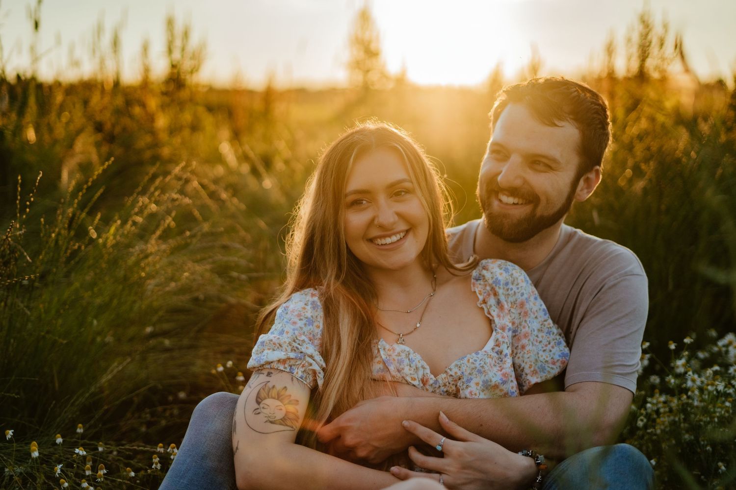 Couple sitting in golden field at sunset, woman smiling in floral dress while man embraces her from behind.
