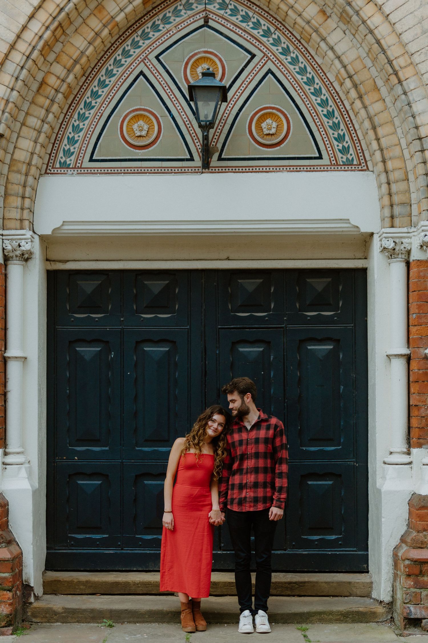 Couple holding hands and smiling in front of ornate arched church doorway in matching red outfits.