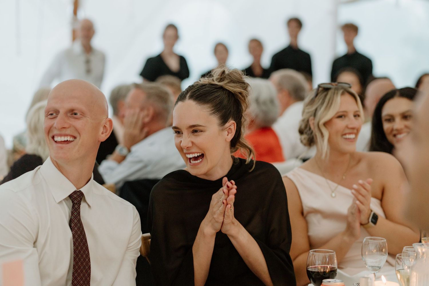 Wedding guests laughing and clapping during reception speeches inside a white tent venue.